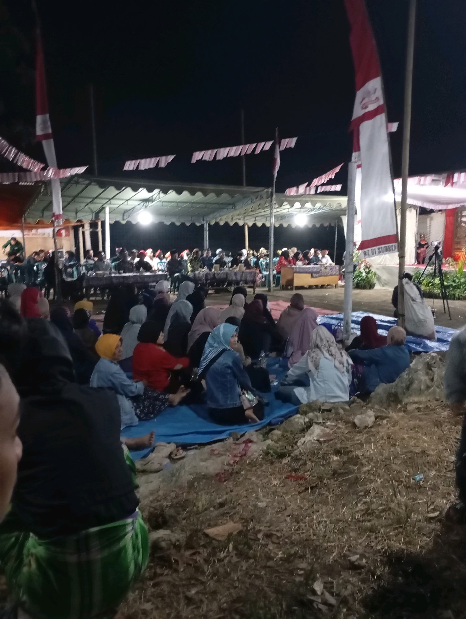 The image depicts an outdoor gathering at night in Ketepung, Indonesia. A large group of people are seated on blue tarps spread on the ground, facing a raised platform or stage. Many individuals are wearing headscarves, suggesting a community event, possibly religious or cultural. A tent structure with lighting is visible in the background, illuminating the area. Indonesian flags are displayed prominently, one of which is a tall red and white flag. The ground is uneven and covered with dry grass and dirt. In the foreground, a person wearing a green and white checkered sarong is partially visible. The overall scene conveys a sense of communal assembly under the night sky.