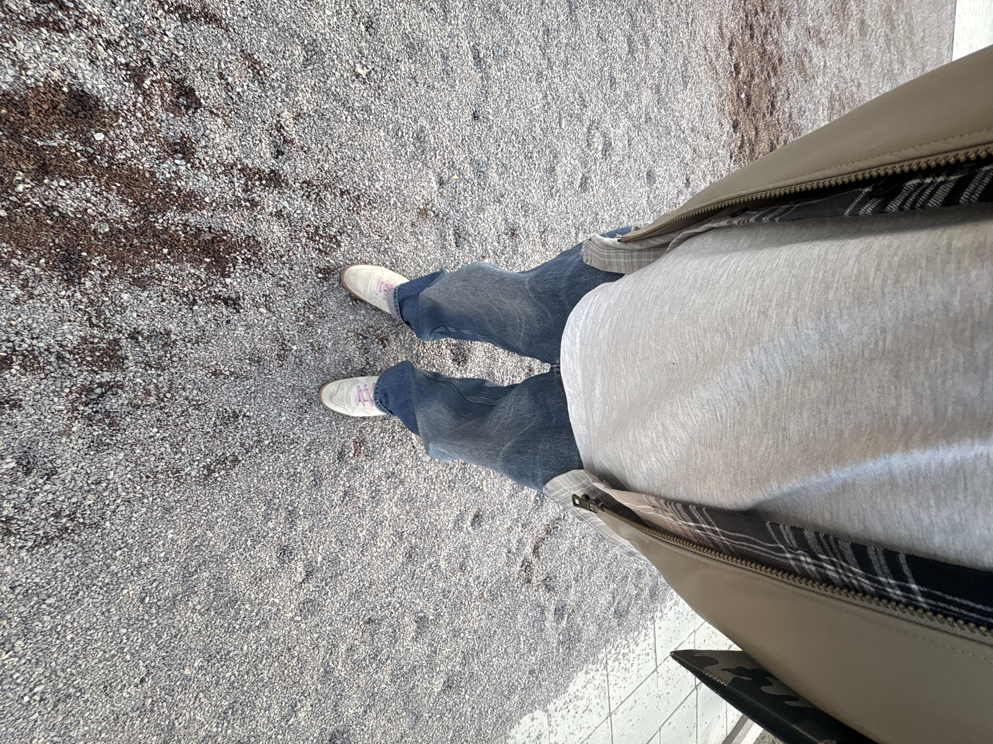 A medium-high angle shot depicts a person standing on a gravel surface, likely a sidewalk or path. The person is wearing blue jeans, off-white sneakers with pink laces, and a light grey long-sleeved shirt under a beige jacket with a visible black and white plaid lining. The gravel surface appears to be a mix of light and dark grey stones. In the lower right corner, a small section of white tiled flooring is visible. The location cannot be determined with the given information to be Moscow, Russia.