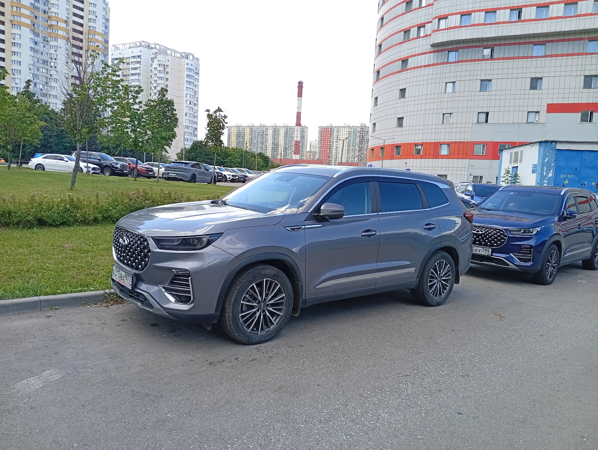 In the city of Unknown, Unknown, a grey Chery Tiggo 8 Pro is parked on an asphalt surface. The car is facing right, with its front bumper and grill clearly visible. To the right of the Chery, a blue car, also parked, is partially visible. In the background, several high-rise apartment buildings are lined up, and a factory chimney with red and white stripes stands tall. Trees and green grass are present in the middle ground, creating a contrast between the urban environment and nature. The sky is overcast.