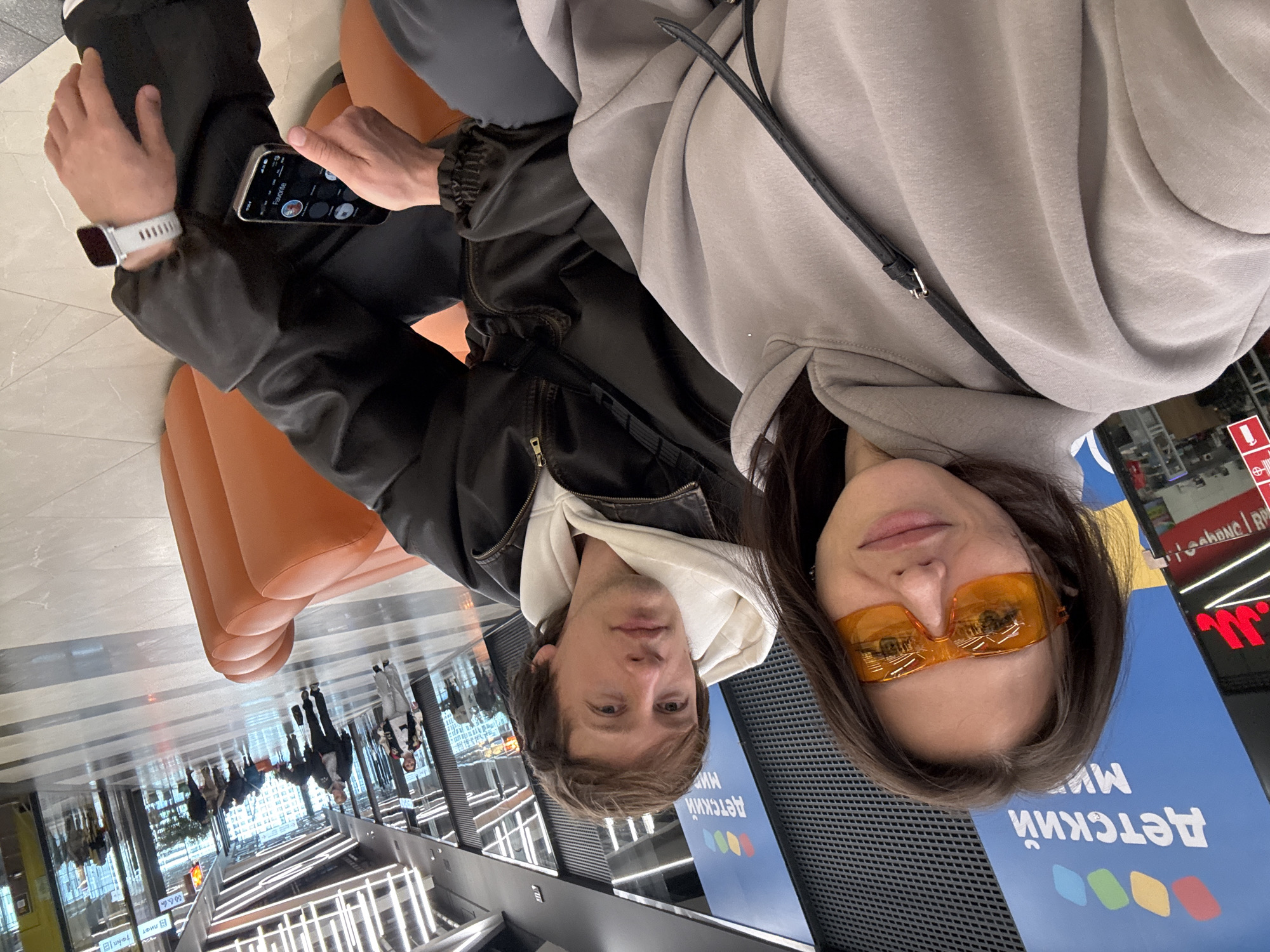 Two individuals are seated on an orange bench inside what appears to be a shopping mall or large commercial space, located in Murino, Russia. The woman is wearing orange-tinted glasses and a light gray hoodie with a black shoulder bag strap across her chest. The man next to her is wearing a black leather jacket over a white hooded sweatshirt. He is holding a smartphone, and a smartwatch is visible on his wrist. The floor reflects the overhead lighting fixtures and pedestrian traffic. In the background is a sign written in Russian, suggesting the location is in Russia.
