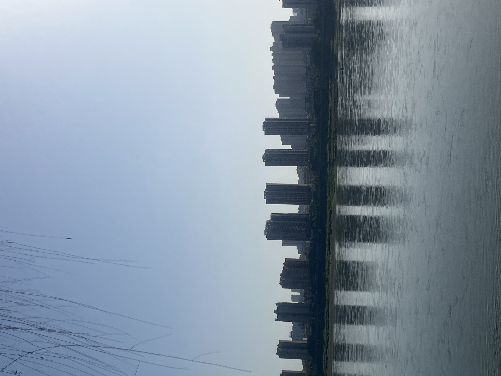 The image captures a panoramic view of a modern cityscape situated along a wide river under a bright, hazy sky. The scene is divided horizontally into three main sections.
The leftmost portion of the frame is dominated by a light, almost white sky that gradually transitions to a pale blue towards the bottom. In the lower left corner, a tangle of thin, dark, out-of-focus branches or wires from a foreground tree extends diagonally upwards, creating a delicate pattern against the sky. A small, dark, indistinct object is faintly visible in the upper left sky area.
The central part of the image features a long line of densely packed, tall buildings that form the city's skyline along the riverbank. These high-rise structures appear silhouetted and somewhat indistinct due to the atmospheric haze, showcasing various shades of grey and dark tones. A narrow strip of dark green vegetation, likely trees or grass, runs between the base of the buildings and the river's edge.
The rightmost section of the image is occupied by the wide river, whose surface reflects the light from the sky and the dark forms of the buildings. These reflections create prominent vertical streaks of light and shadow across the water, giving it a textured, almost shimmering appearance. The river's surface shows subtle ripples. A tiny, dark boat or object is barely discernible on the river, adding a minute point of interest.
The light quality suggests it is daytime, possibly a slightly overcast or hazy day, contributing to the soft, muted tones throughout the image. There are no people clearly visible, and the scene conveys a sense of quiet urban expanse. No visible text is present. The location context provided is Jiangjin, China.