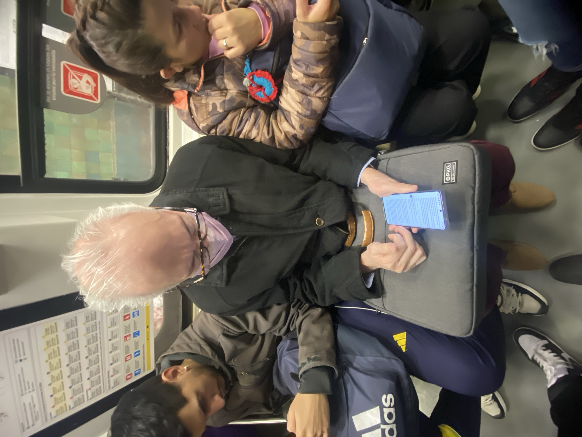 The image depicts an interior view, likely a subway car in Buenos Aires, Argentina, judging by the route map affixed to the window. Three individuals are visible, seated closely. A woman with brown hair and a patterned jacket sits nearest the window, partially obscured. An older man with white hair and a face mask sits next to her, holding a blue smartphone and a gray backpack. A younger man with dark hair, wearing a brown jacket, is seated by the older man. A navy blue Adidas backpack can be seen between the two men. Other people's feet are visible in the background. The overall environment is crowded, suggesting a busy public transit setting.