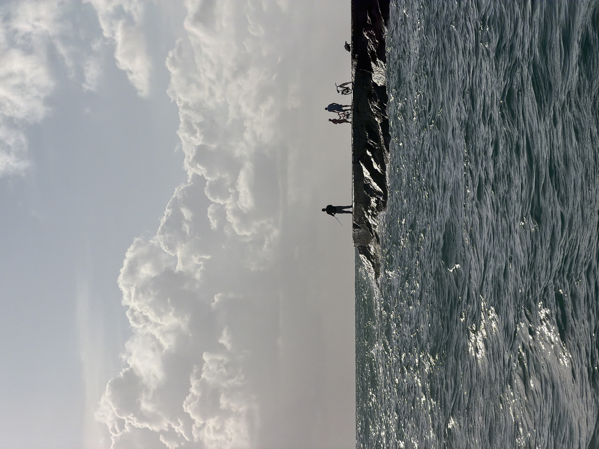 Here is a forensic description of the image:
The image depicts a scene in Bal Harbour Village, Florida. A breakwater structure divides the frame, with the ocean on the right and a cloudy sky on the left. Several individuals are silhouetted against the sky on the breakwater. One person is fishing at the end, while a group with bicycles stands further back. The ocean exhibits wave action and a rippled surface, reflecting light. The sky features prominent cumulus clouds.