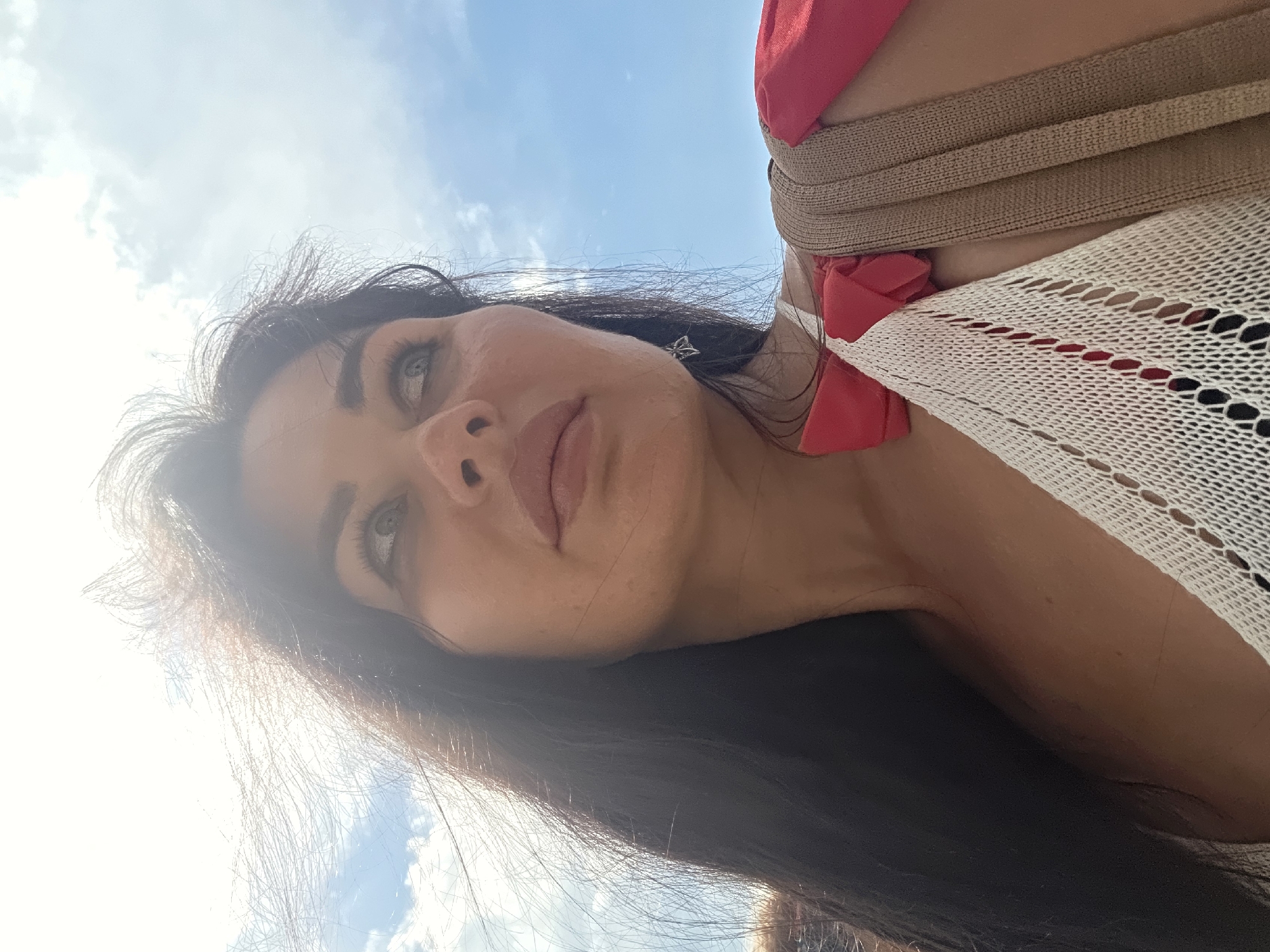 A low-angle, close-up, outdoor, self-portrait photograph captures a woman's face and upper torso against a bright blue sky with scattered clouds, strongly suggesting a location in Sochi, Russia, known for its sunny climate and coastal views. The woman, with long, dark, wind-blown hair, gazes upwards and to her right, her expression thoughtful. Her features are clearly defined, with prominent cheekbones and full lips. She wears a white, crocheted top with a bright red bow detail, and a tan strap, likely from a bag or dress, crosses her shoulder. The lighting is harsh, with the sun creating a lens flare effect and highlighting strands of her hair. The image is focused on her face and shoulders, with the sky occupying the upper half of the frame.