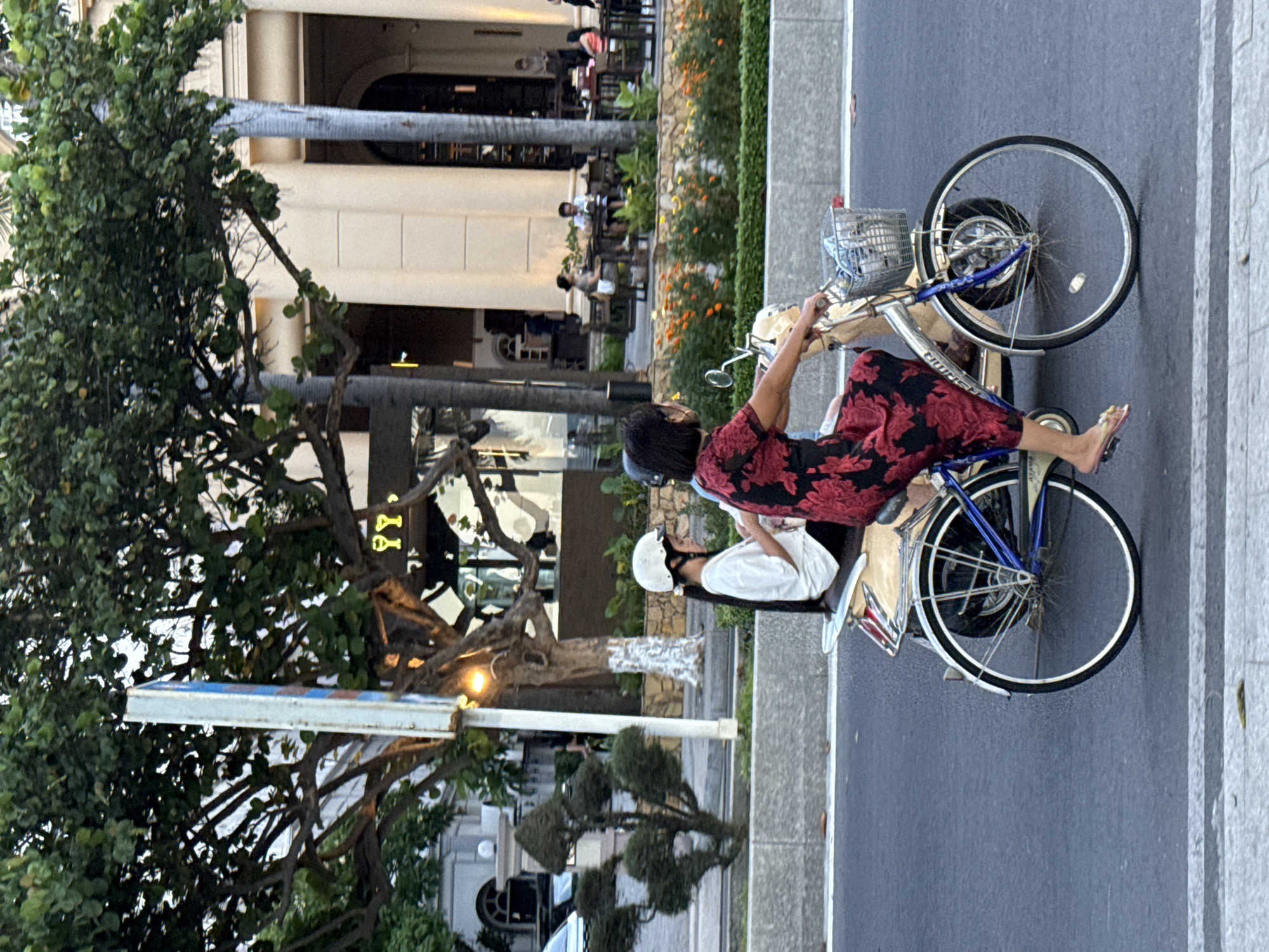 This image depicts two people on bicycles on a street in Nha Trang Ward, Vietnam. In the foreground, a person wearing a red and black floral dress and a helmet is riding a bicycle. The bicycle has a basket on the front and appears to be a vintage model. Behind this person, another individual wearing a white shirt and a helmet is also on a bicycle.
The scene appears to be an urban environment with a building in the background that has large windows and an outdoor seating area with tables and chairs, suggesting a cafe or restaurant. There are trees with green foliage and some flowering plants along the side of the street. The lighting suggests it could be daytime, possibly late afternoon or early morning, given the soft light. The road is asphalt, and there's a sidewalk made of stone. The overall atmosphere seems casual and everyday. There is no visible text in the image.