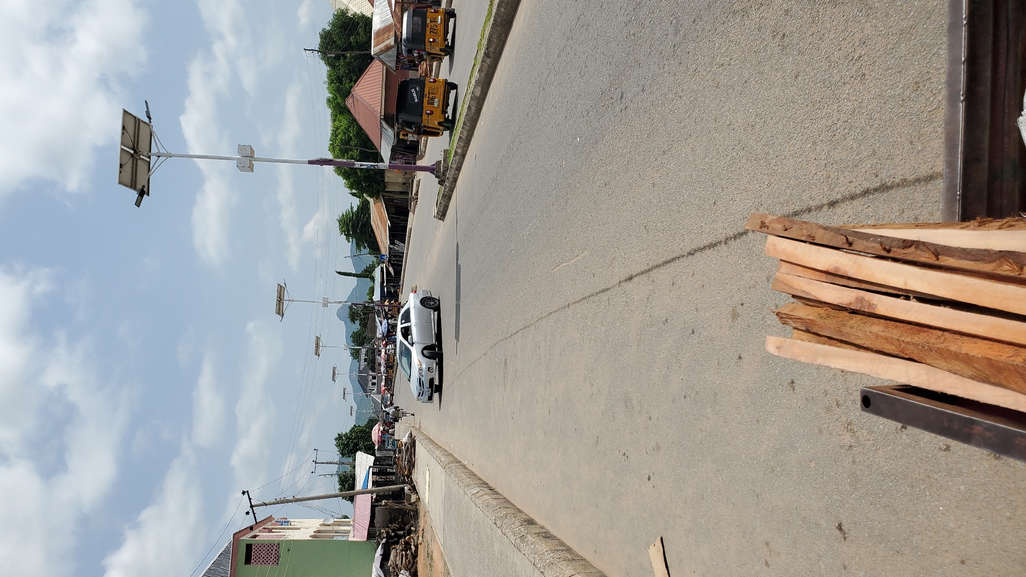 The image depicts a street scene in Jalingo, Nigeria, likely a commercial or residential area. In the foreground, a pile of rough-cut lumber is visible. The paved road runs through the middle of the frame, with a concrete sidewalk on the left. Several solar-powered streetlights are positioned along the road. On the left side of the road, a light green building with pink trim and multiple windows is present, with shops or stalls in front of it. A white sedan is driving on the road. Further down the road, two yellow three-wheeled vehicles, commonly known as Keke Napep, are also in motion. Buildings with corrugated iron roofs line the right side of the street, and trees are scattered throughout the scene. In the distance, mountains are visible under a partly cloudy sky. The overall impression is of a typical day in a Nigerian town, with economic activity and infrastructure present.
