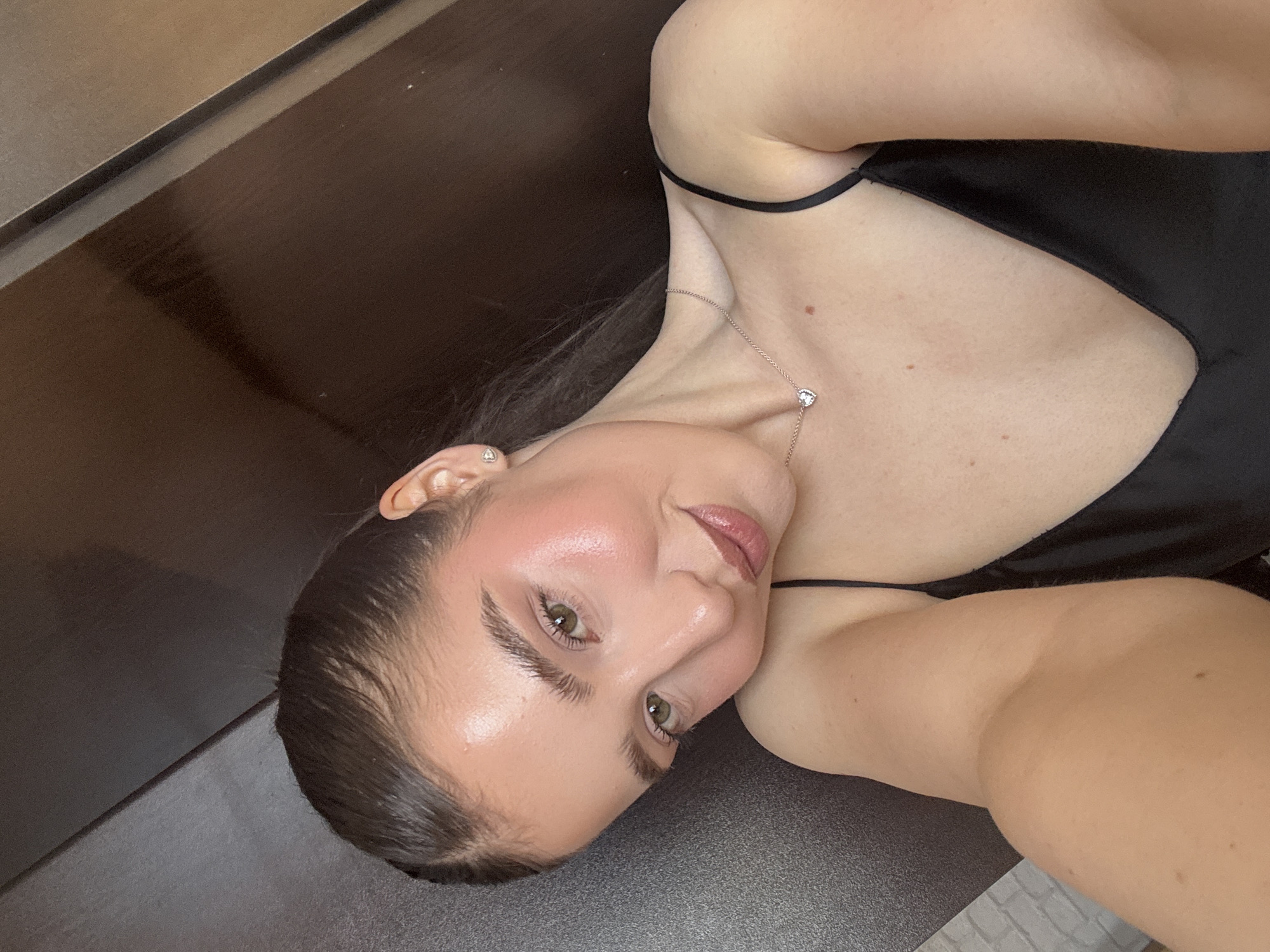 A woman with fair skin and dark hair is lying on a dark surface, possibly a table or floor. She is wearing a black camisole-style top with thin straps and a delicate silver necklace with a small pendant. Her face is tilted toward the camera, and she has light green eyes, defined eyebrows, and full lips. The lighting highlights her cheekbones. The location appears to be indoors.