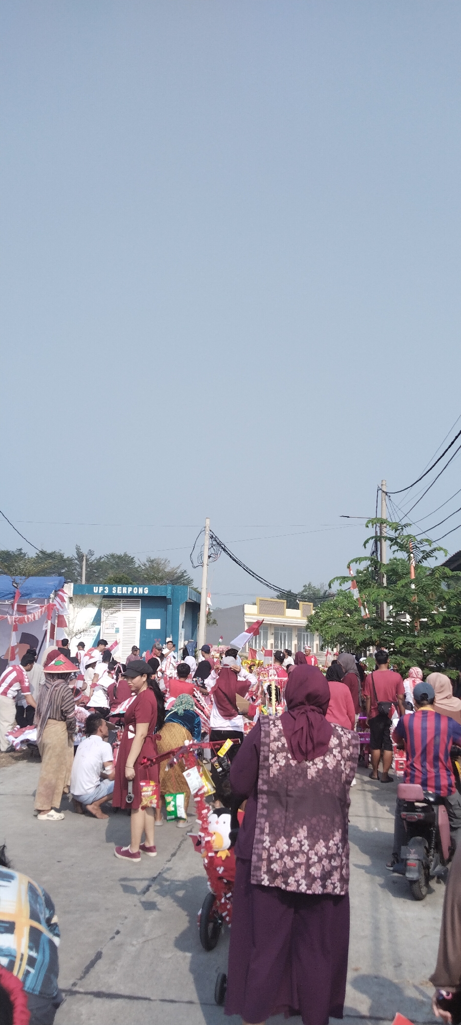 The image depicts an outdoor gathering of people in Taban, Indonesia. The sky is clear and blue. In the background, there is a building with the sign "UP3 SERPONG". Many people are dressed in red and white, suggesting a celebratory event, possibly related to Indonesian Independence Day. There are flags and decorations in red and white throughout the scene. Several people are walking, and some are gathered around stalls or displays. One person is riding a motorcycle. The ground appears to be paved. There are trees and electrical poles visible. The overall atmosphere seems festive and communal.