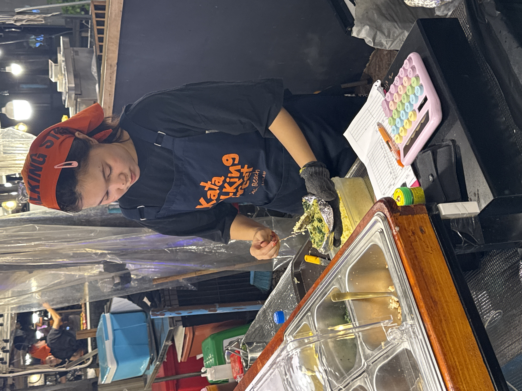 The image shows a woman preparing food at a street food stall in Karon, Thailand. She is wearing a black t-shirt, a dark blue apron, and an orange bandana with "WALKING STREET" printed on it. She is also wearing a black glove on her right hand. The woman is focused on her task, her head bowed as she works with some food wrapped in foil.
In the background, there are various elements of a street food setup. We can see cooking equipment, plastic sheeting used as a barrier, and bright lights, suggesting it is evening or night. A pink calculator with colorful buttons is visible on the counter, along with a paper with writing on it and a pen. There are also stainless steel containers for ingredients or prepared food, and a wooden counter. Other people and stalls are faintly visible in the background, indicating a busy market environment. The overall scene depicts a typical street food operation, likely in a tourist area given the "Walking Street" text.