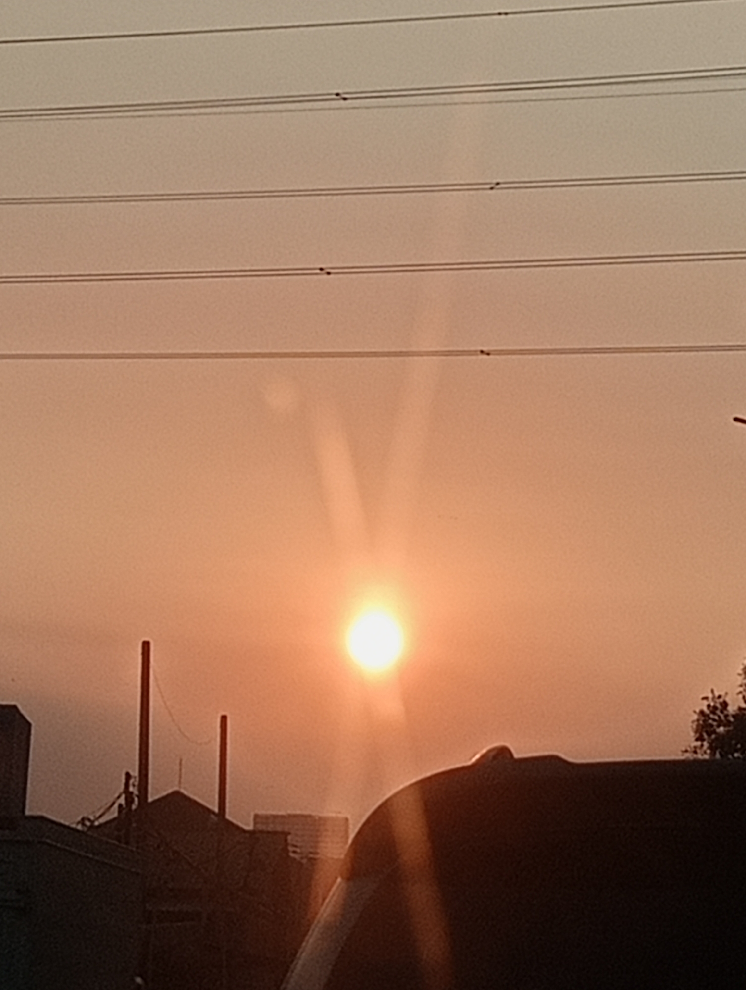 The image shows a sunset over Surabaya, Indonesia. The sun is a bright orb in the hazy sky, casting a warm, orange glow over the scene. In the foreground, the silhouette of a car can be seen, suggesting the photo was taken from a vehicle. Behind the car, there are the dark shapes of buildings and structures, some with visible utility poles and wires stretching across the frame. The overall atmosphere is one of a typical urban sunset, possibly captured during twilight hours.