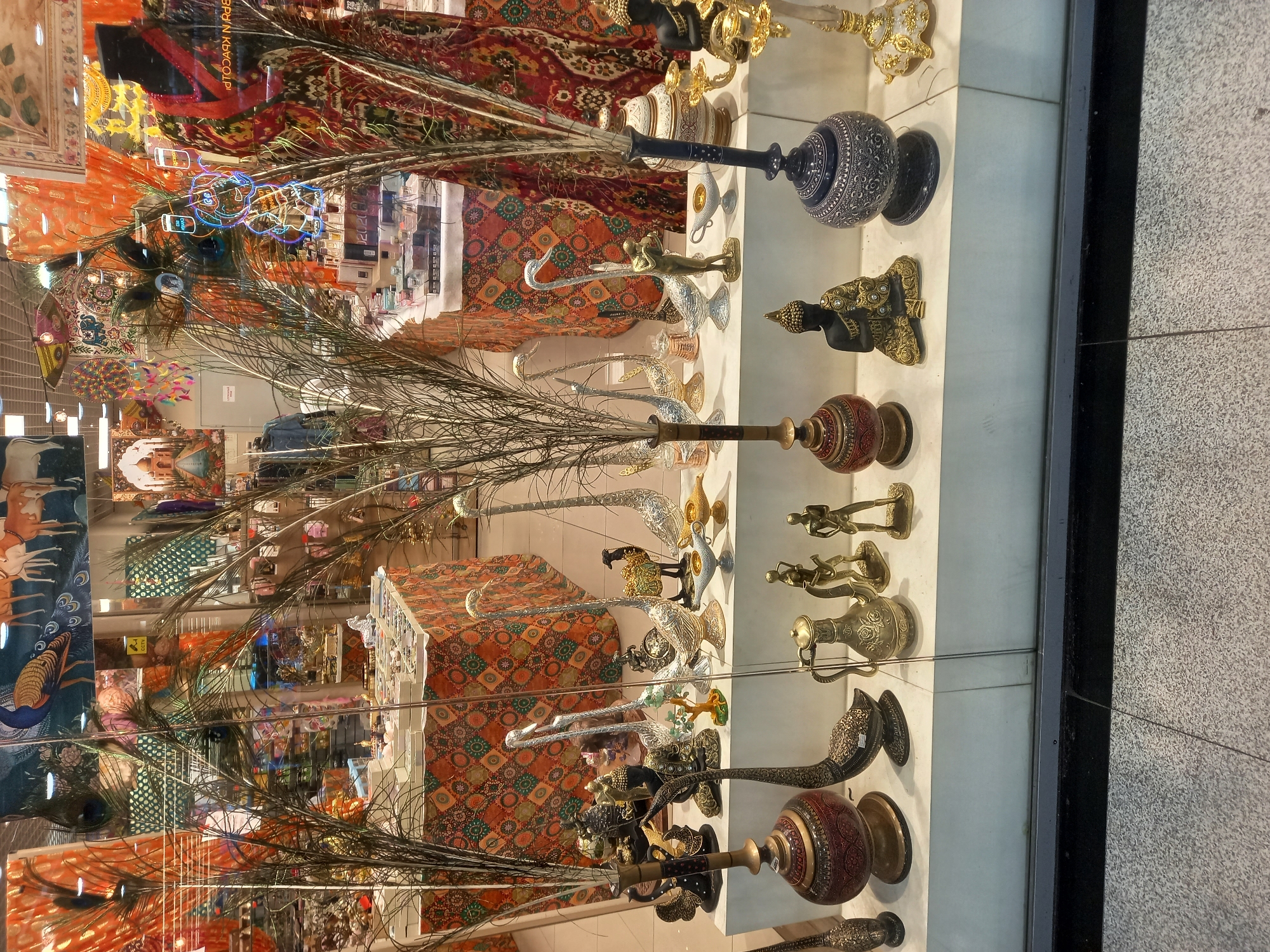 This image shows a display window of a shop, likely in Yekaterinburg, Russia, featuring various decorative items. The foreground is dominated by a collection of metal figurines and vases. These include several ornate brass-colored vases with intricate carvings, some shaped like swans or geese, and a dark blue decorative vase with white patterns. There are also small figurines of people and a seated Buddha statue. In the middle ground, more decorative pieces are displayed, including silver-colored swan figurines, a camel figurine, and oil lamps. Tall, slender vases with dried reeds and peacock feathers are positioned prominently in front of the merchandise. The background reveals the interior of the shop, with colorful fabrics, tapestries depicting animals and Indian architecture, and shelves stocked with various items. The overall impression is of an ethnic or artisanal goods store.
