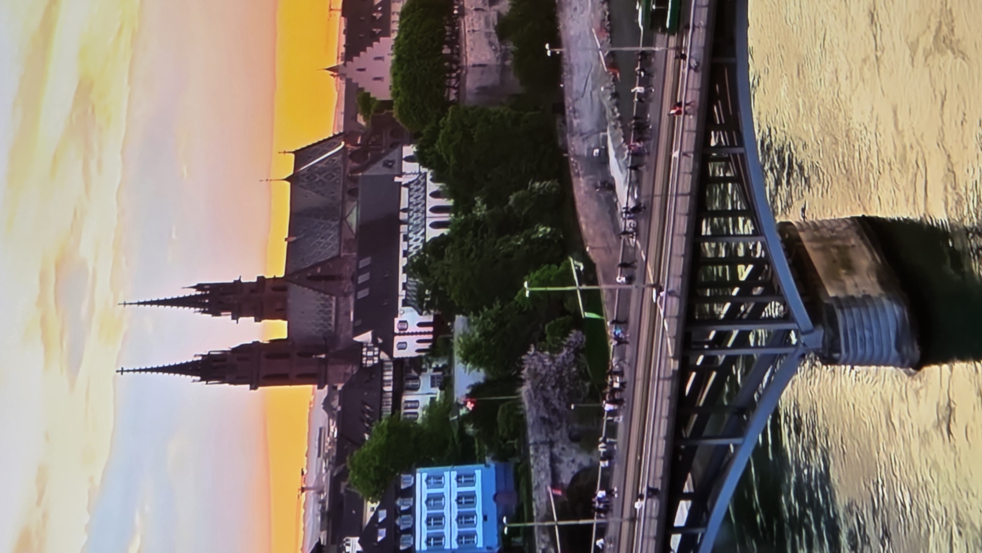An aerial view captures a city skyline at sunset, with a prominent cathedral featuring twin spires in the background. A bridge spans a body of water in the foreground, carrying traffic and pedestrians. Buildings of various architectural styles, including a blue facade structure with multiple windows, are visible along the riverbank, interspersed with lush green trees. The sky is a gradient of orange, yellow, and light blue, indicative of twilight. There is no indication that this location is in Batumi, Georgia.