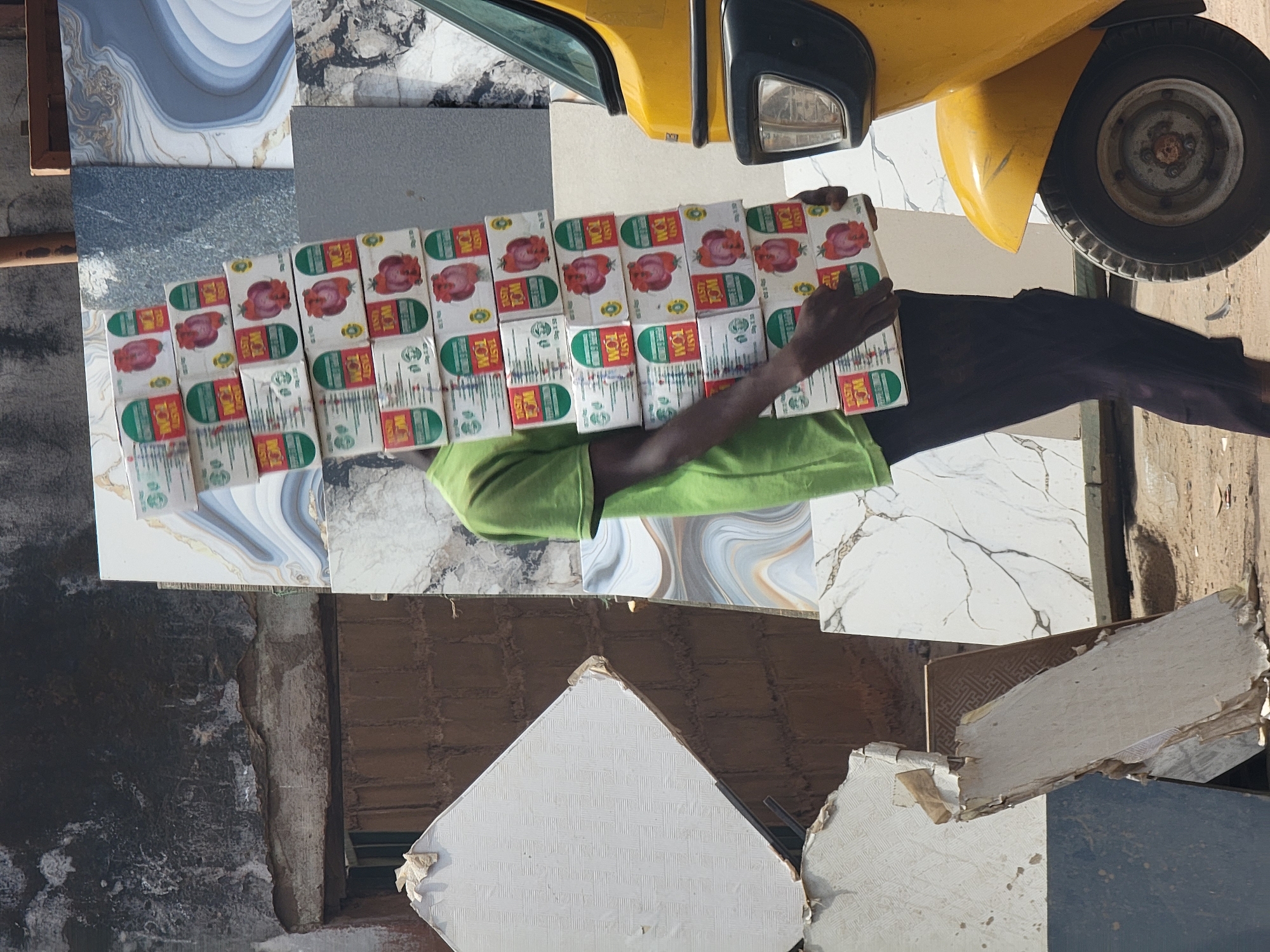 A person in Maiburiji, Nigeria, wearing a green shirt and dark pants, carries multiple boxes of "Tasty Tom" brand tomato paste near a yellow vehicle, possibly a taxi or auto-rickshaw. The background includes various tiles and building materials, suggesting an urban or commercial environment.