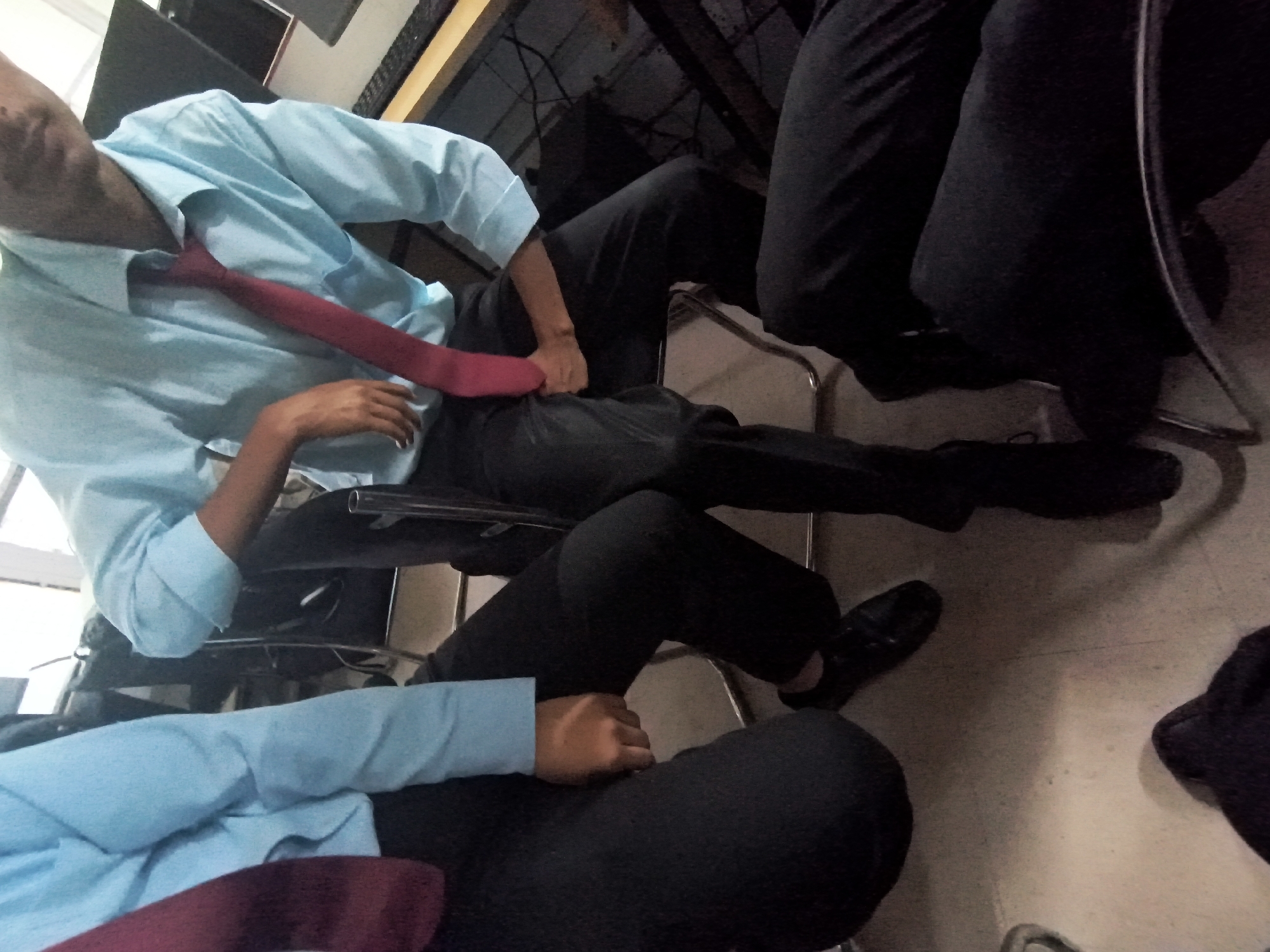 In a classroom or office environment, likely within Dharamshala, India, two individuals are captured in a seated position. The foreground shows the legs and lower torsos of two people. One person, on the left, wears black trousers and a maroon tie, with a light blue shirt partially visible. Their left leg is bent, with a black shoe visible. The other person, to the right, also wears black trousers and a light blue shirt, with a maroon tie. Their legs are crossed, and one hand is visible resting on their thigh. In the background, another person in a light blue shirt and maroon tie is seated, leaning back in a chair, with a partial view of their face. A desk with a keyboard and monitor is visible in the far background. The floor appears to be tiled or linoleum. The overall lighting suggests an indoor setting, possibly during the daytime.
