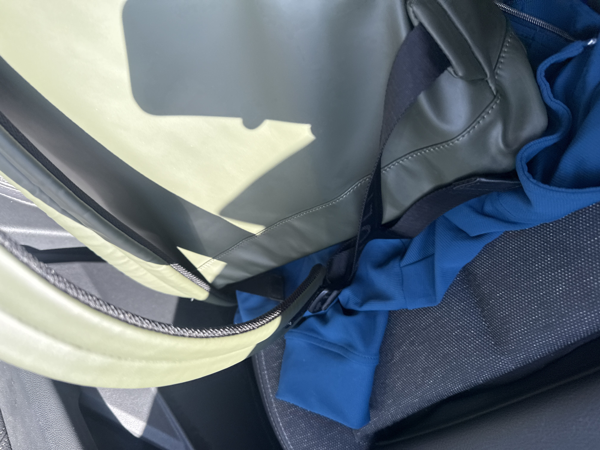 The image presents a close-up, slightly angled view inside a vehicle's interior. A prominent, large olive-green or light-khaki colored bag, possibly a duffel bag or backpack, occupies the left and central parts of the frame. It features a smooth, possibly synthetic material with visible stitching and black trim along its edges and straps, one of which includes a black buckle.
Direct sunlight from above casts distinct, sharp shadows across the bag's surface and the surrounding area, indicating a bright, sunny daytime environment. Tucked against and partially beneath the bag is a bright royal blue garment, likely a long-sleeved shirt or jacket, resting on the vehicle's dark gray seat upholstery. The upholstery has a finely textured, almost mesh-like pattern. On the far left, a section of black plastic or rubber trim from the car's interior is visible.
The scene suggests items are being transported or temporarily stored within a car during the day. No specific activity is depicted, nor are any individuals present. There is no visible text in the image. The context places this scene in Puebla, Mexico.