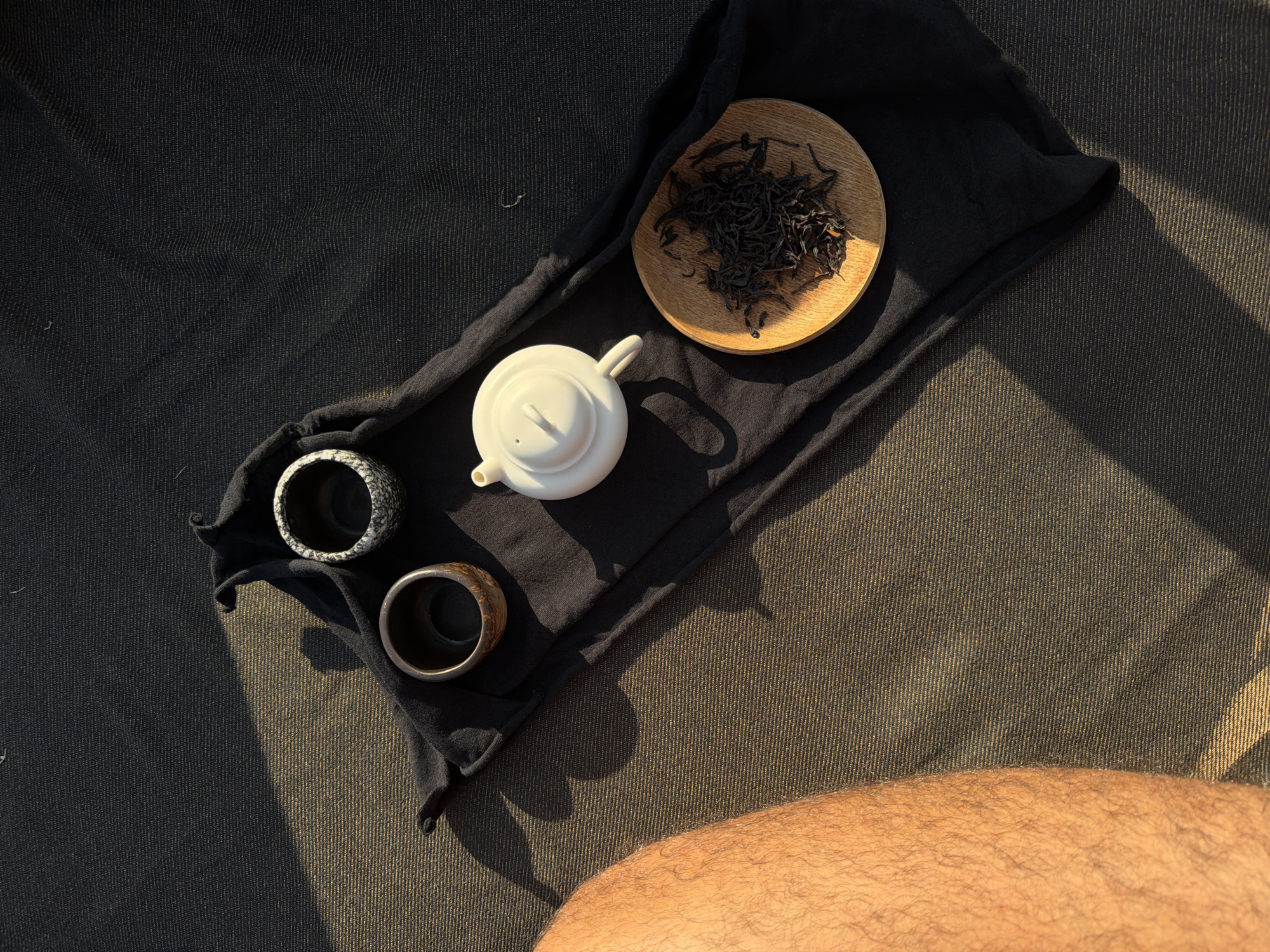 An overhead view captures a tea ceremony arrangement on a dark, textured surface, situated in Selemadeg, Indonesia. A black fabric runner spans horizontally across the frame. Positioned on the runner are a white ceramic teapot, a dark brown wooden tea cup, and a speckled grey ceramic tea cup. A small, light-colored wooden plate holding dark, dried tea leaves is also on the runner. Strong, directional sunlight illuminates the setup from the upper right, creating distinct shadows on the dark background. The underlying surface exhibits a fine, dark, ribbed textile pattern. The upper section of a bare, tanned human leg with visible hair occupies the left side of the image, receiving direct illumination.