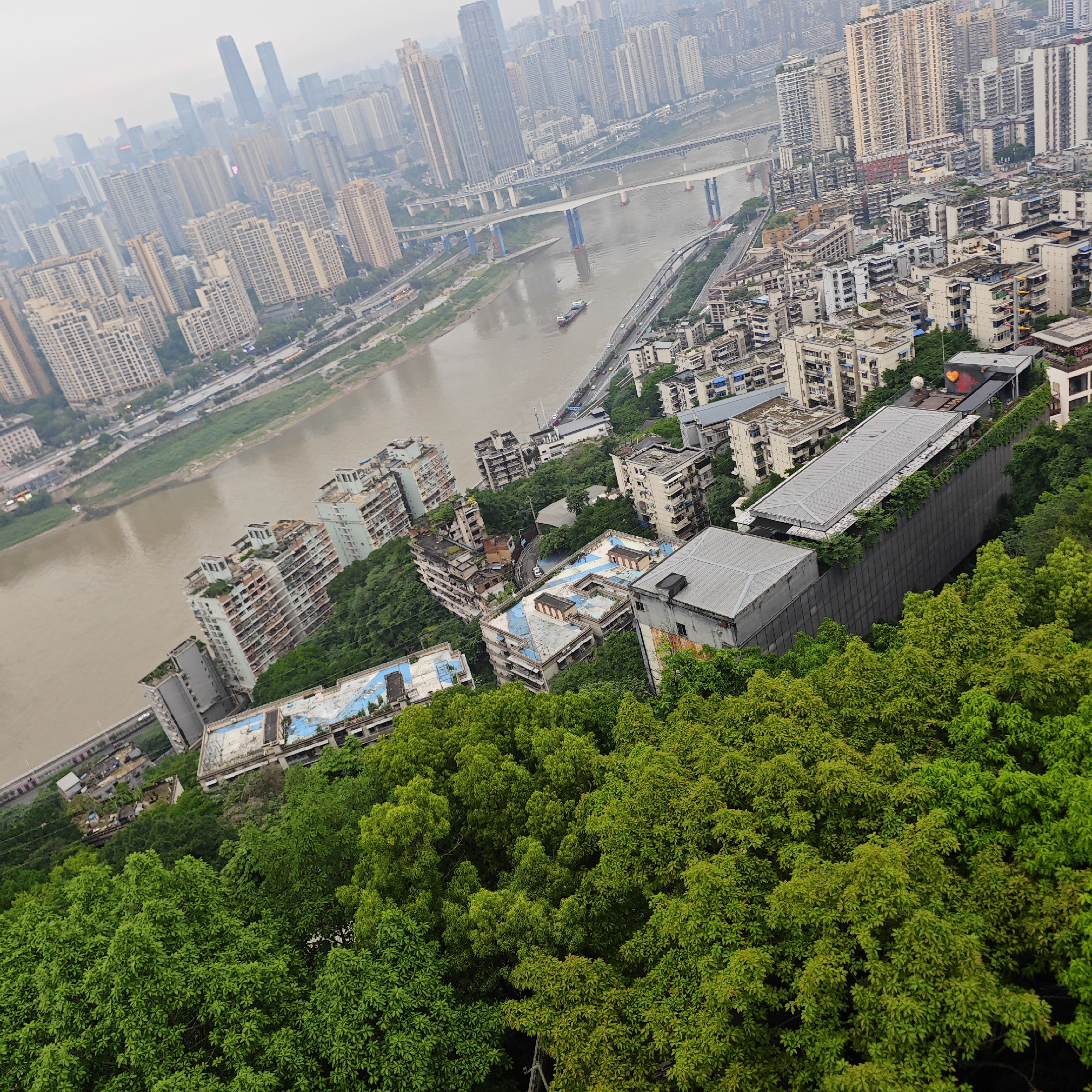 The image presents an elevated view in Yuzhong District, China, showcasing a landscape of urban development interspersed with natural elements. In the foreground, dense green foliage dominates, suggesting a hillside or elevated park area. Behind the foliage, there are several mid-rise buildings with rooftop structures, including some with flat, gray rooftops and others with deteriorating blue-painted roofs. A long, gray concrete wall with greenery growing along the top appears to support some of the buildings.
A brown river flows through the scene, bisecting the urban area. A cargo ship is visible on the water. Several bridges span the river, carrying vehicular traffic. On both sides of the river, there are roads and paths running along the banks, adjacent to the river's edge.
Beyond the river, the urban landscape consists of numerous high-rise residential and commercial buildings, indicative of a densely populated urban center. The buildings vary in height and architectural style, creating a diverse skyline. The background of the image is hazy, which partially obscures the distant structures.