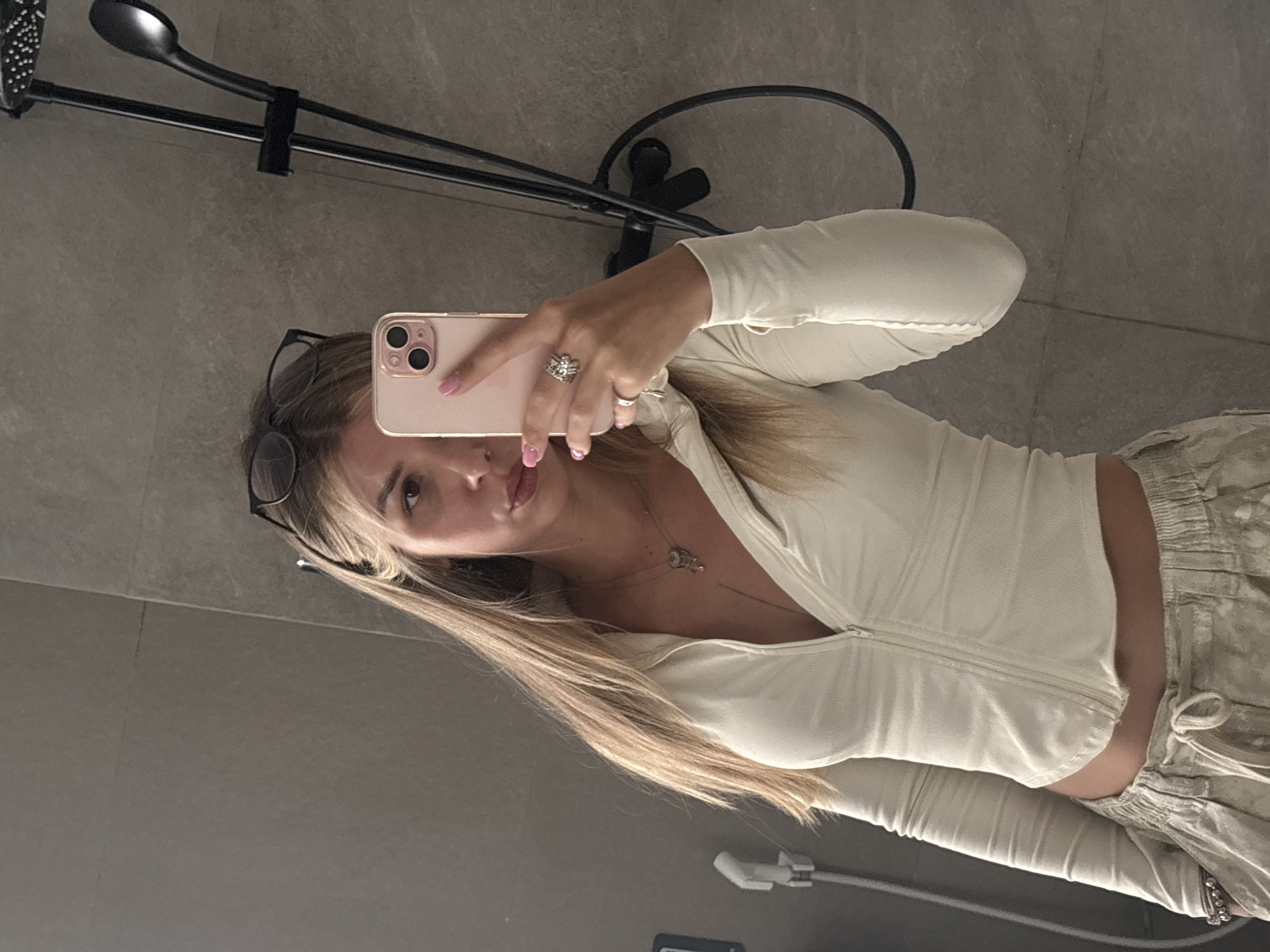 A young woman with blonde hair and glasses is taking a selfie in a bathroom. She is wearing a white zip-up top and beige pants. She is holding a pink iPhone in her right hand and is wearing rings on her fingers. The bathroom has grey tiled walls and a black showerhead is visible in the background.