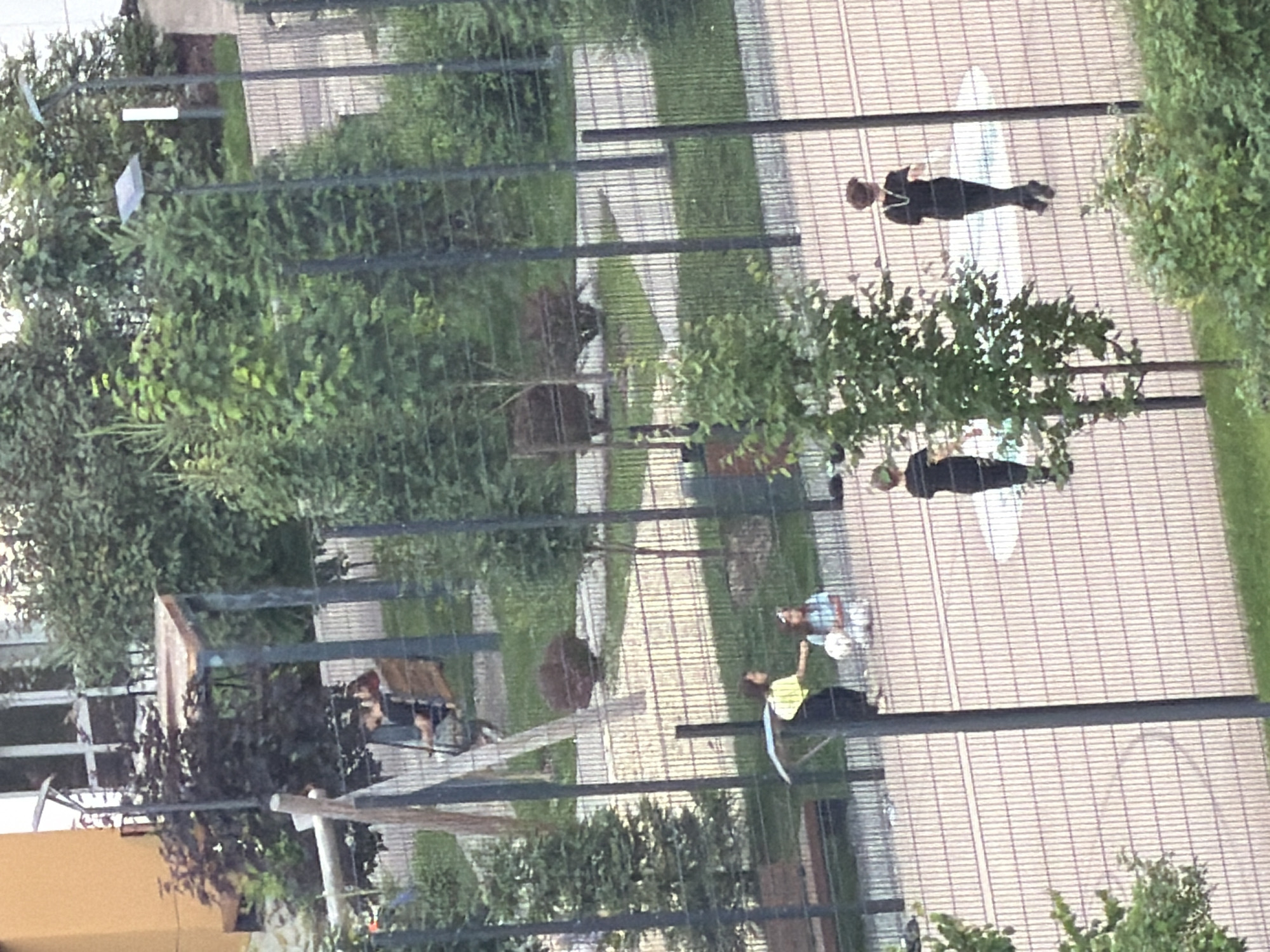 The image captures an outdoor scene through a fence, likely in Kazan, Russia. Several people are visible, engaged in various activities. In the upper part of the frame, a person in black clothing stands on a paved area. Further down, another person in a black outfit is also standing, while two individuals are seated. One seated person wears a yellow shirt and black pants, and the other is in a blue and white striped top with white pants. They appear to be interacting, with one person holding a white object, possibly a ball. The setting includes lush green trees and bushes, suggesting a park or an outdoor recreational area. The paved ground is marked with lines, creating a grid pattern. The presence of trees and the general ambiance suggest it is daytime. There is no visible text in the image.