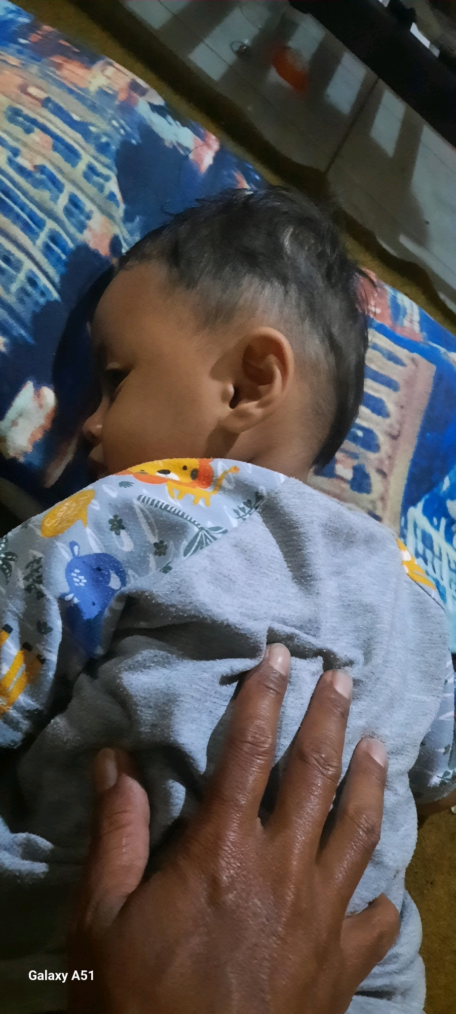 A close-up, over-the-shoulder view of a sleeping baby in Jatitengah, Indonesia, with a hand resting on the baby's back. The baby is lying on a blue and white patterned blanket. The baby is wearing a grey onesie with cartoon animal prints, including lions and elephants. The hand, which appears to be an adult's, is a darker skin tone and has a few visible wrinkles. The background is blurred, but suggests an indoor setting with a floor and possibly furniture. The Samsung Galaxy A51 watermark is visible in the bottom left corner.