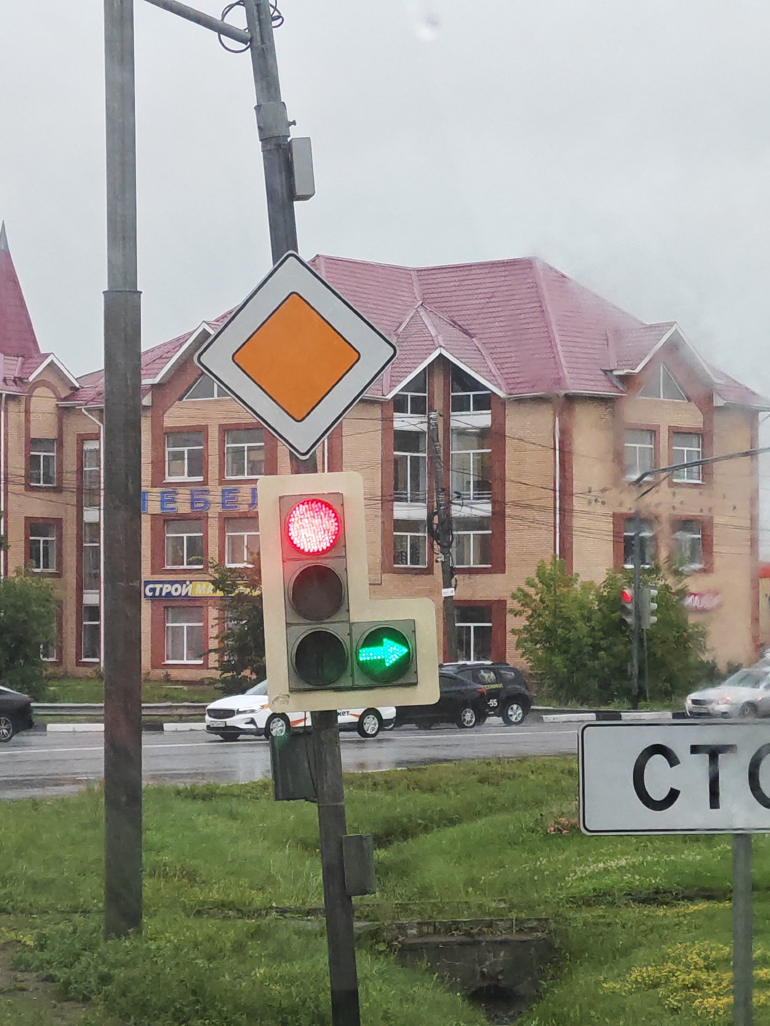 The image shows a street scene in Lukhovitsy, Russia, on a cloudy day. A traffic light is prominently featured, displaying a red light at the top and a green arrow pointing right at the bottom. The traffic light is mounted on a pole, which also supports a diamond-shaped sign with an orange center, indicating a priority road.
In the background, a multi-story brick building with a red roof is visible. The building has numerous windows and signs. One sign on the building reads "СТРОЙ М."
Several cars are present on the wet road. A white car is stopped at the intersection, and a black SUV is driving away from the camera. Another white car is visible further down the road, also moving.
The foreground is filled with green grass. A street sign with the letters "СТО" is visible on the right side of the image. The overall impression is of a typical urban intersection during a period of rain.