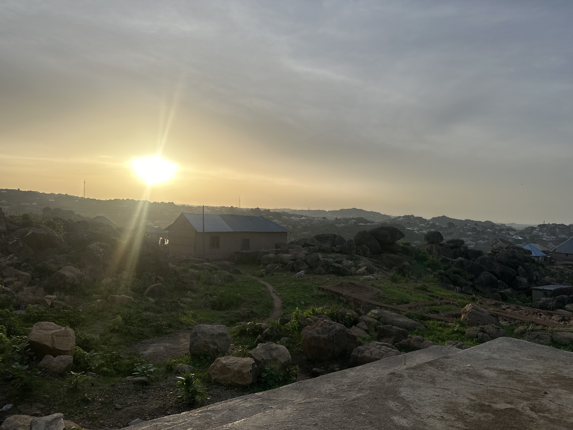 This media file captures a wide landscape view in Jos, Nigeria, likely during early morning or late afternoon. The dominant subjects are the expansive, hazy sky with a bright, low-lying sun, and a rugged, rocky terrain.
In the foreground and middle ground, numerous large, grey and brown boulders are scattered across a gently sloping, green, and partially barren landscape. Patches of green grass and small shrubs grow between the rocks, with a narrow dirt path winding through. A small, simple, single-story house with a light blue or grey corrugated metal roof and a light brown/beige exterior stands prominently in the middle ground, surrounded by rocks and vegetation. A faint, small white animal, possibly a sheep or goat, is visible near the house in the distance.
The sun, positioned on the left side of the frame, is a powerful source of light, creating a strong lens flare that radiates across the sky and illuminates parts of the rocky ground. The sky is largely overcast or hazy, with lighter, yellowish tones near the sun gradually transitioning to a muted grey-blue further away.
In the far distance, beyond the immediate rocky hills, a sprawling settlement or town can be seen, characterized by numerous houses with varying roof colors. The overall scene conveys a sense of natural ruggedness combined with human habitation. There is no explicit activity taking place, and no visible text. The notable details include the striking sun glare, the characteristic rocky landscape of Jos, and the hazy atmospheric conditions contributing to the soft, diffused light.