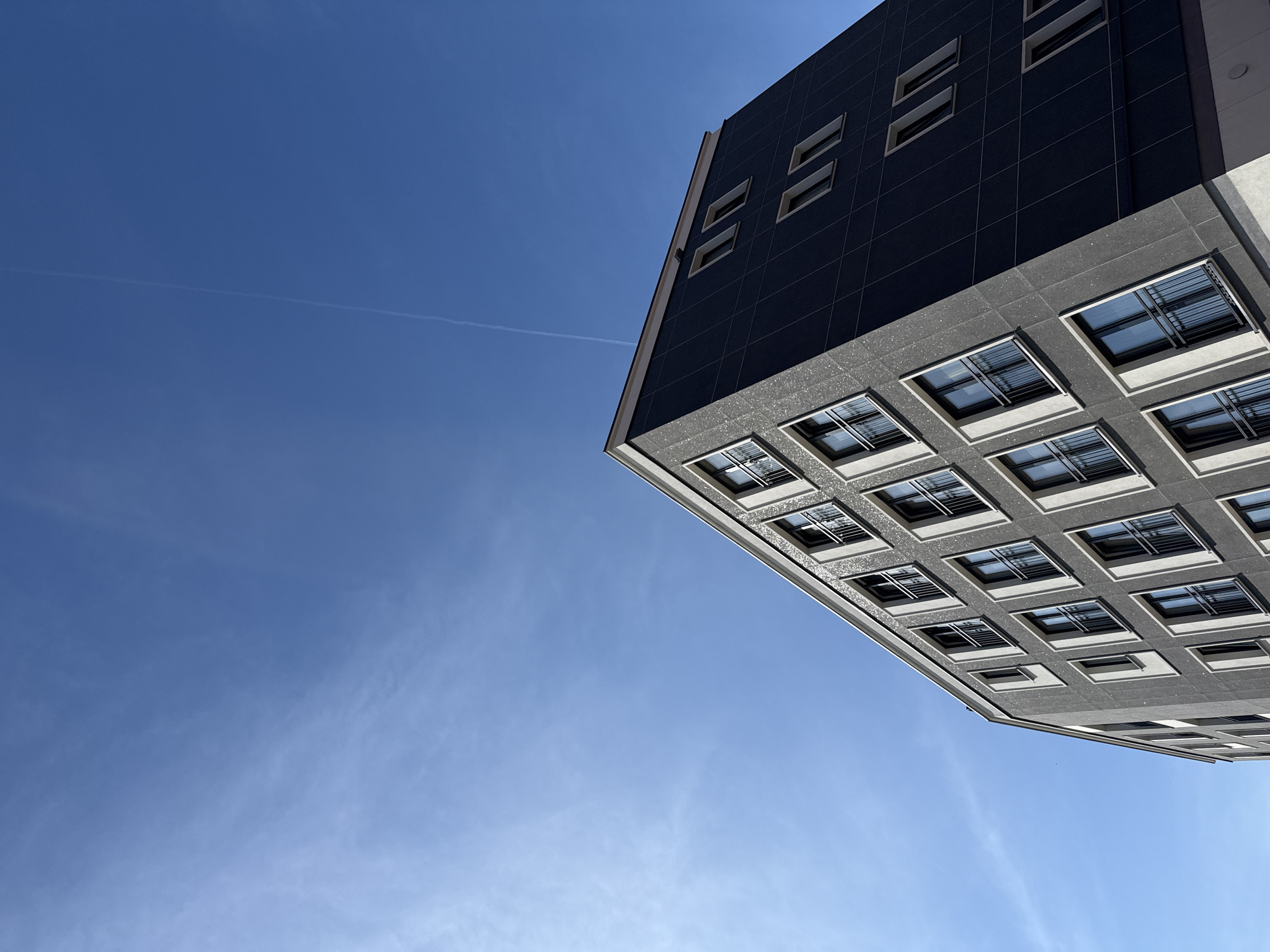 A modern, grey-clad building with white-trimmed windows is viewed from below against a clear blue sky. The building's angled facade emphasizes its geometric structure. A faint contrail from an aircraft stretches diagonally across the sky. The image is cropped to focus on the upper portion of the building and the sky.