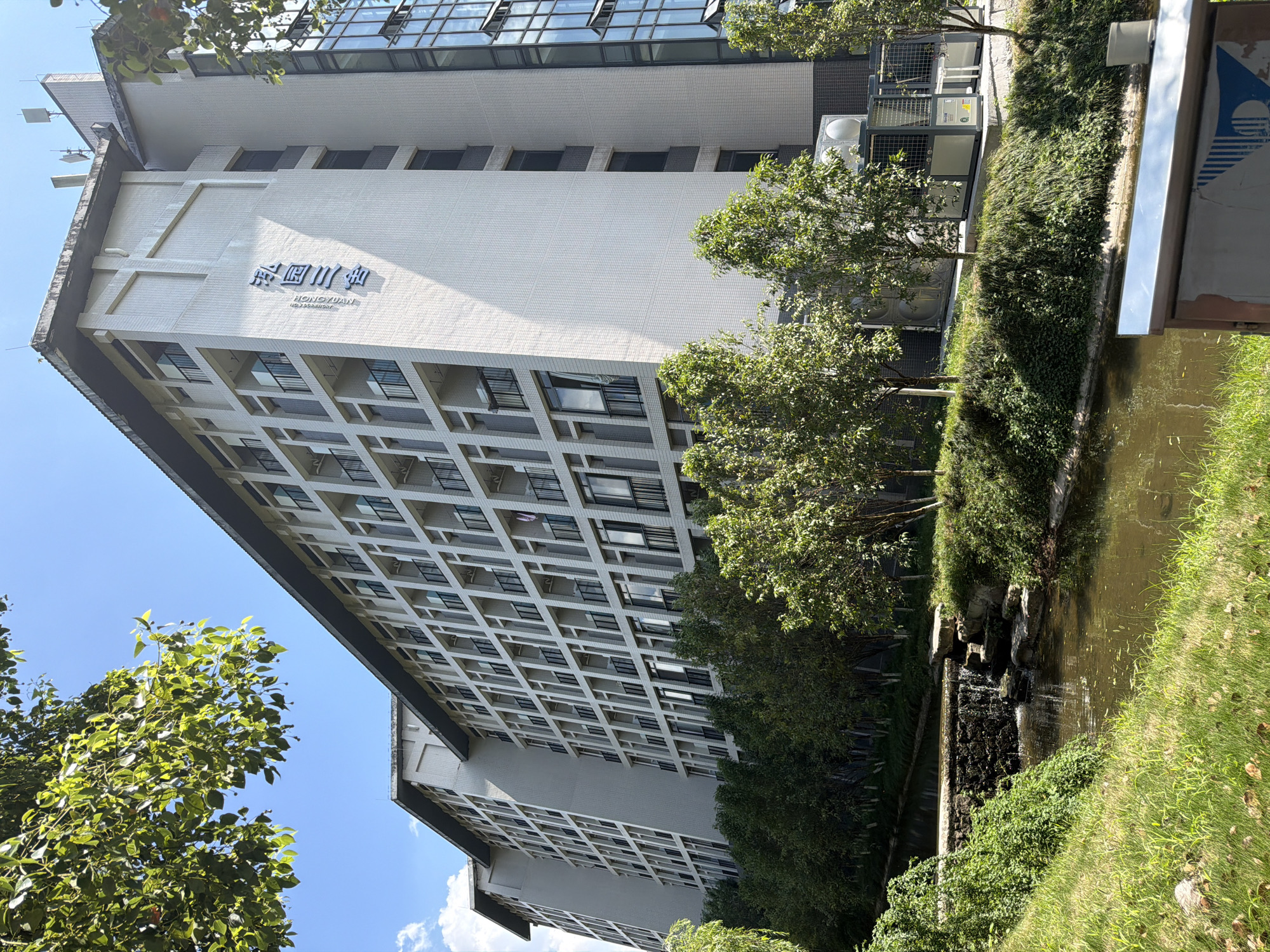 In the city of Jiangjin, China, a long, multi-story building with a light-colored tiled facade stands under a clear blue sky. The building has numerous windows and balconies with black railings, arranged in a grid pattern. The upper portion of the building features a dark-colored roofline. On the upper facade, large Chinese characters are visible, along with smaller English text. To the left of the building, a body of water with a small waterfall cascades over rocks. Lush green trees and grass surround the water and the building. In the foreground, there is a metal railing with some visible markings. The scene appears to be a well-maintained campus or residential area.