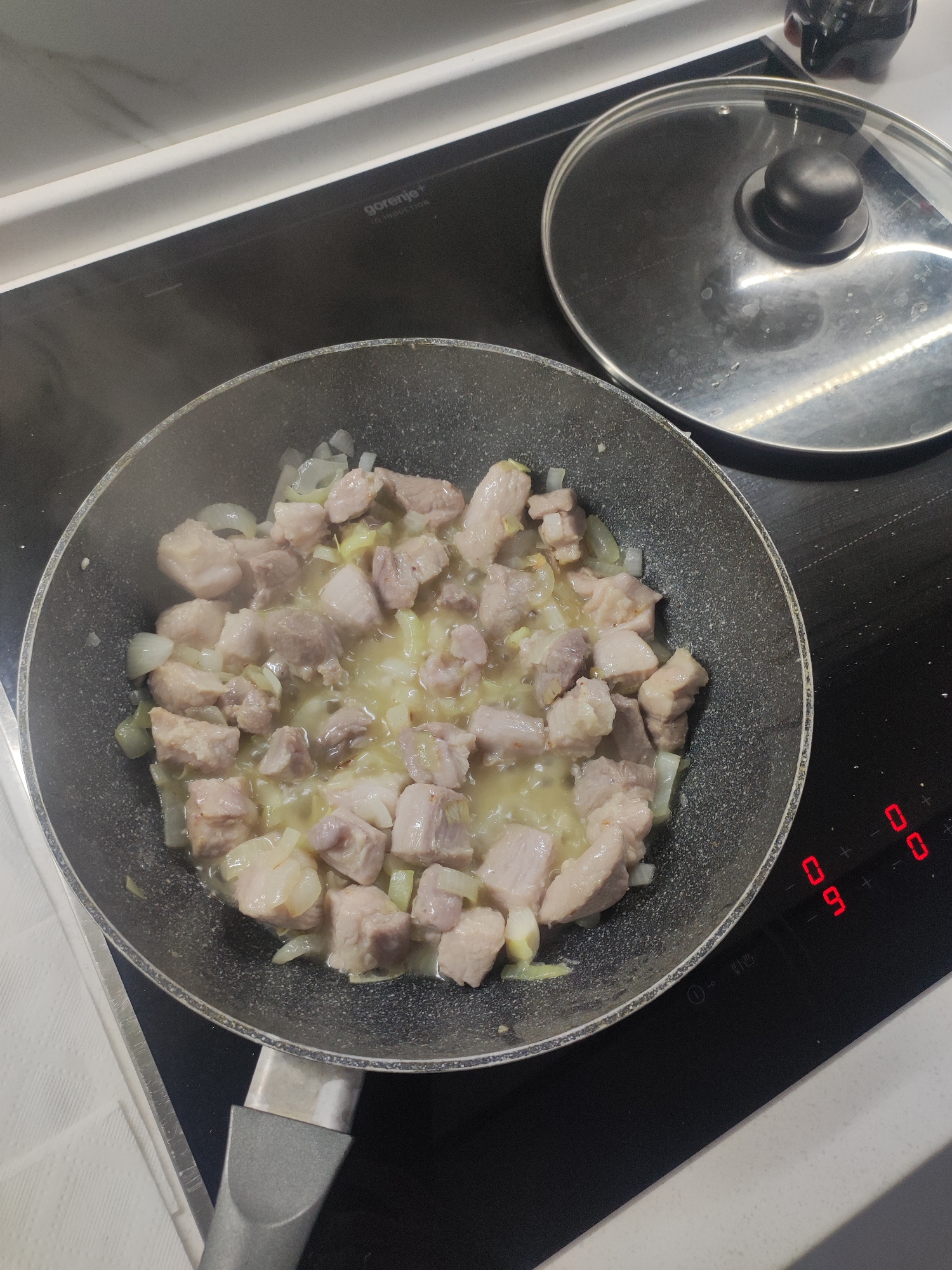 In a Moscow kitchen, pieces of raw pork and chopped onions are being fried in a black non-stick pan on a black induction stovetop. The food is releasing steam, indicating it is actively cooking. A glass lid with a black knob rests on the stovetop next to the pan, suggesting it might have been used recently or is waiting to be placed on the pan. The stovetop has digital controls that are lit up, displaying red numbers. Specifically, "09" is visible on the left, and two "00" indicators are on the right. A white countertop runs along the top edge of the image, and a white paper towel is partially visible on the left. The scene captures a moment of everyday cooking.