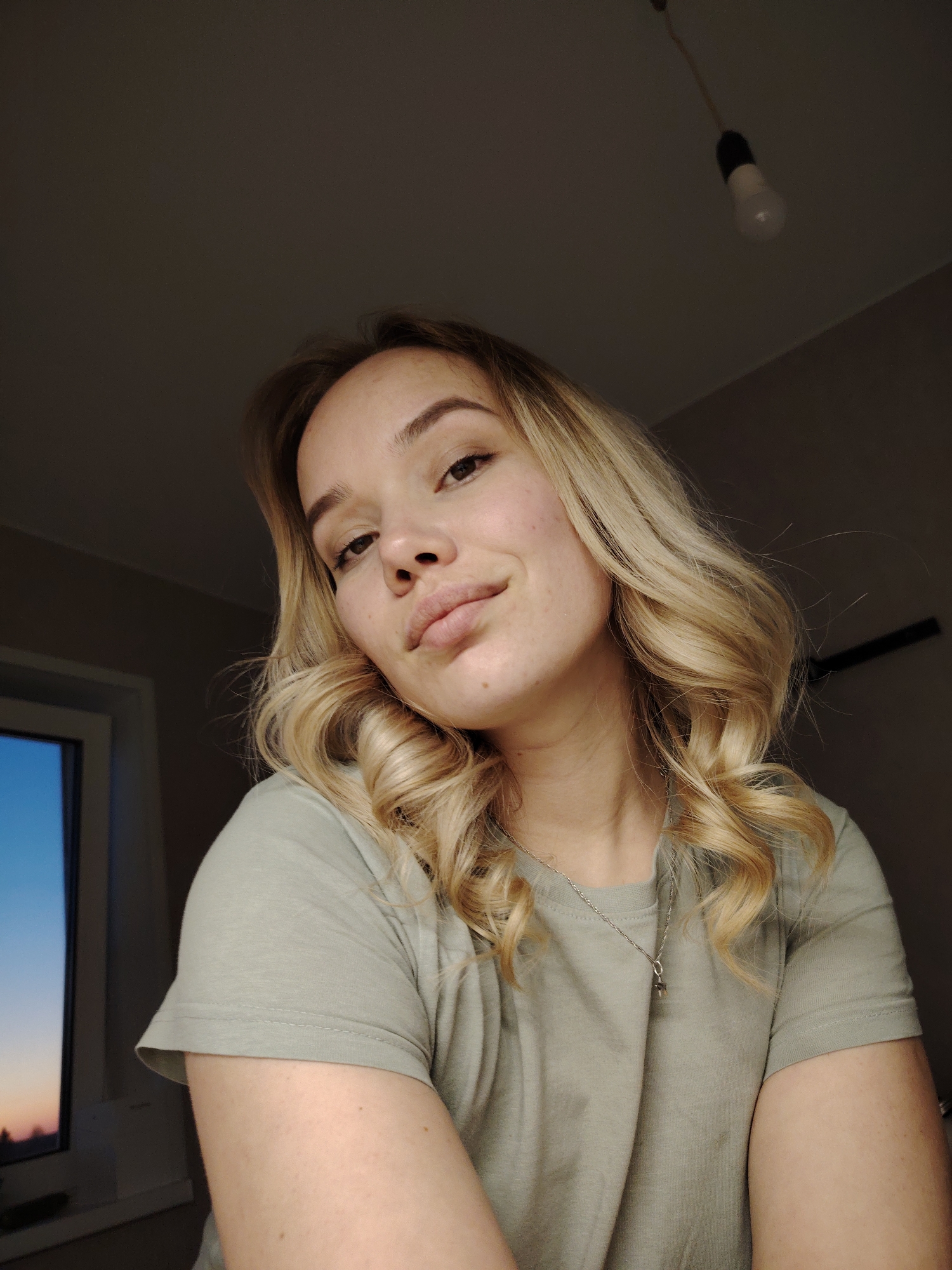 A young woman is posing for a selfie in an interior setting. The lighting is dim, primarily lit from the side with a window showing an evening sky.
The woman has fair skin and light blonde hair styled with curls around her face. She's wearing a light olive green t-shirt and a silver necklace. The room appears to be simply furnished with a bare lightbulb hanging from the ceiling. A window with a view of what appears to be sunset or twilight can be seen to the left.
The location is not verifiable as being in Марковский, Russia based on the visual data.