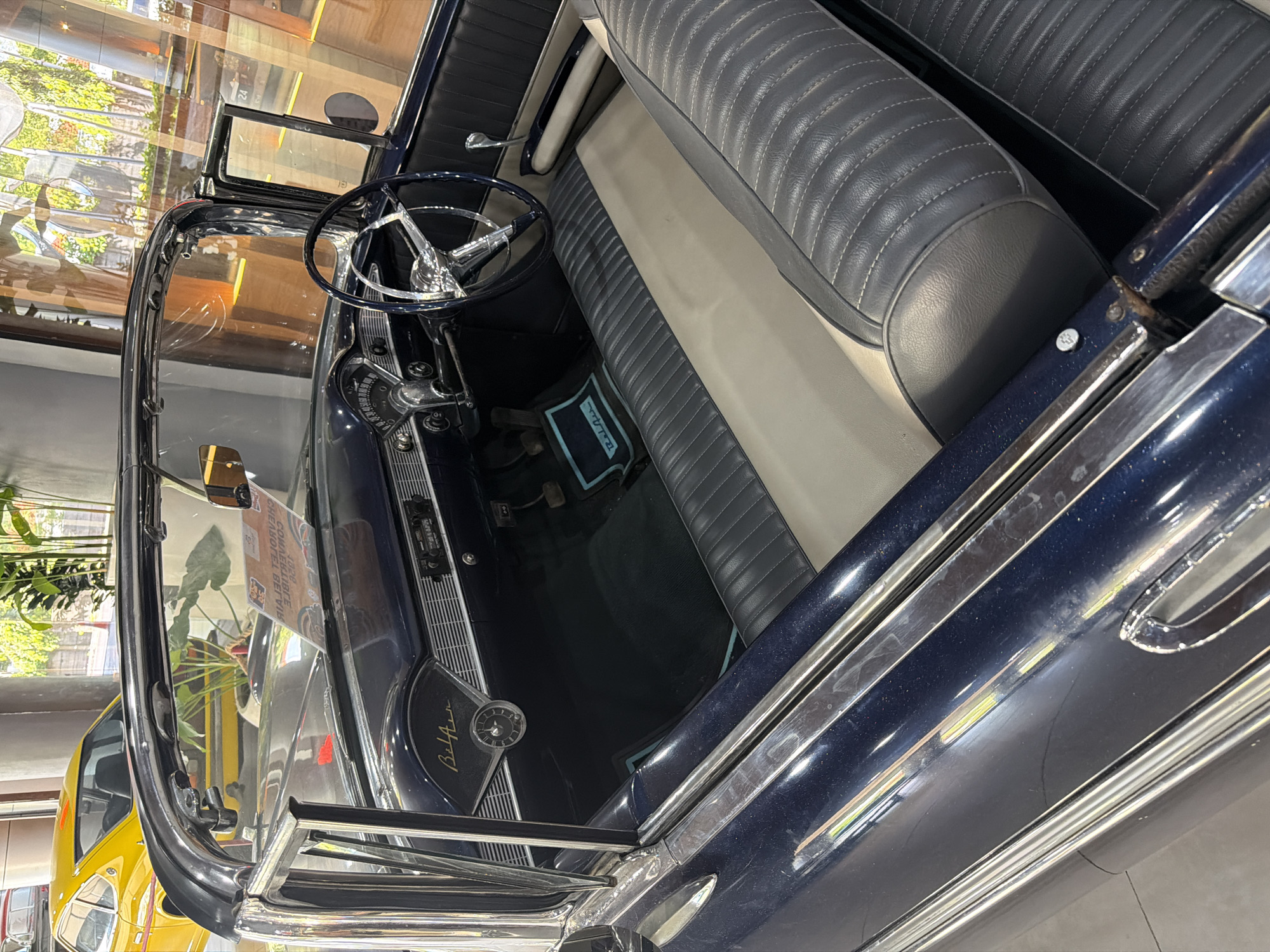 The image captures a detailed, high-angle view of the interior and front passenger side of a vintage dark blue convertible car, likely on display indoors. The car's exterior is a deep, glossy blue, accented with substantial chrome trim around the windshield, windows, and along the side body.
The interior features a two-tone design, with dark blue and lighter gray quilted upholstery on the bench seats. The classic black steering wheel, with chrome spokes, is prominent on the driver's side. The dashboard is equipped with a circular speedometer and other gauges, and the stylized text "Bel Air" is clearly visible on the passenger side, identifying the vehicle as a Chevrolet Bel Air. A dark blue floor mat, accented with light blue, is seen in the footwell.
Various reflections are present across the car's polished surfaces. The windshield reflects an indoor setting with a patterned ceiling and large windows or an open area showcasing bright outdoor light and lush green foliage. A partial reflection of a yellow classic car is visible on the passenger side exterior, implying other vintage vehicles are nearby. A mirrored sign, reflected in the chrome, includes the text "CHEVROLET BEL AIR CONVERTIBLE". The car is stationary, suggesting it is part of an exhibition or collection, in Kuta, Indonesia. No individuals are present in the image.