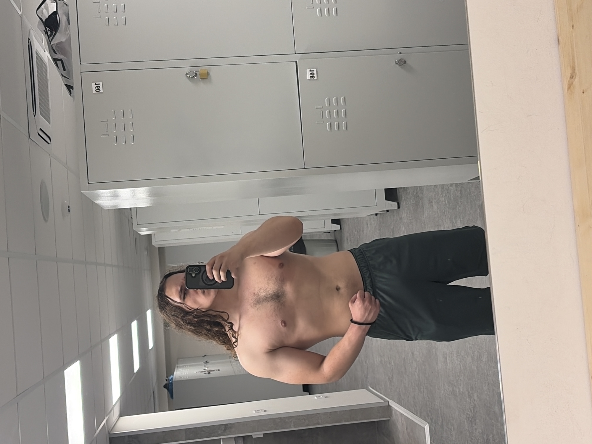 An adult male with long, curly brown hair is captured in a mirror selfie, shirtless and wearing dark green or black athletic shorts. He is holding a black smartphone in his left hand to take the photo. He has a muscular physique, with visible chest hair, and a black wristband on his right wrist.
The setting appears to be a modern and well-lit changing room or locker room, possibly within a gym or sports facility in Plovdiv, Bulgaria. The background features light grey metal lockers with square ventilation patterns, some showing combination locks, including a gold-colored padlock on one. Visible on the lockers are the numbers "801" and "807".
The room has white tiled walls, a light grey floor, and a white counter or vanity in the foreground. Overhead, multiple horizontal fluorescent lights provide bright illumination. In the top left of the reflection, a white bag rests on top of the lockers near what appears to be an air vent or an air conditioning unit. The overall environment is clean and utilitarian. The man's expression is neutral, focused on taking the picture.