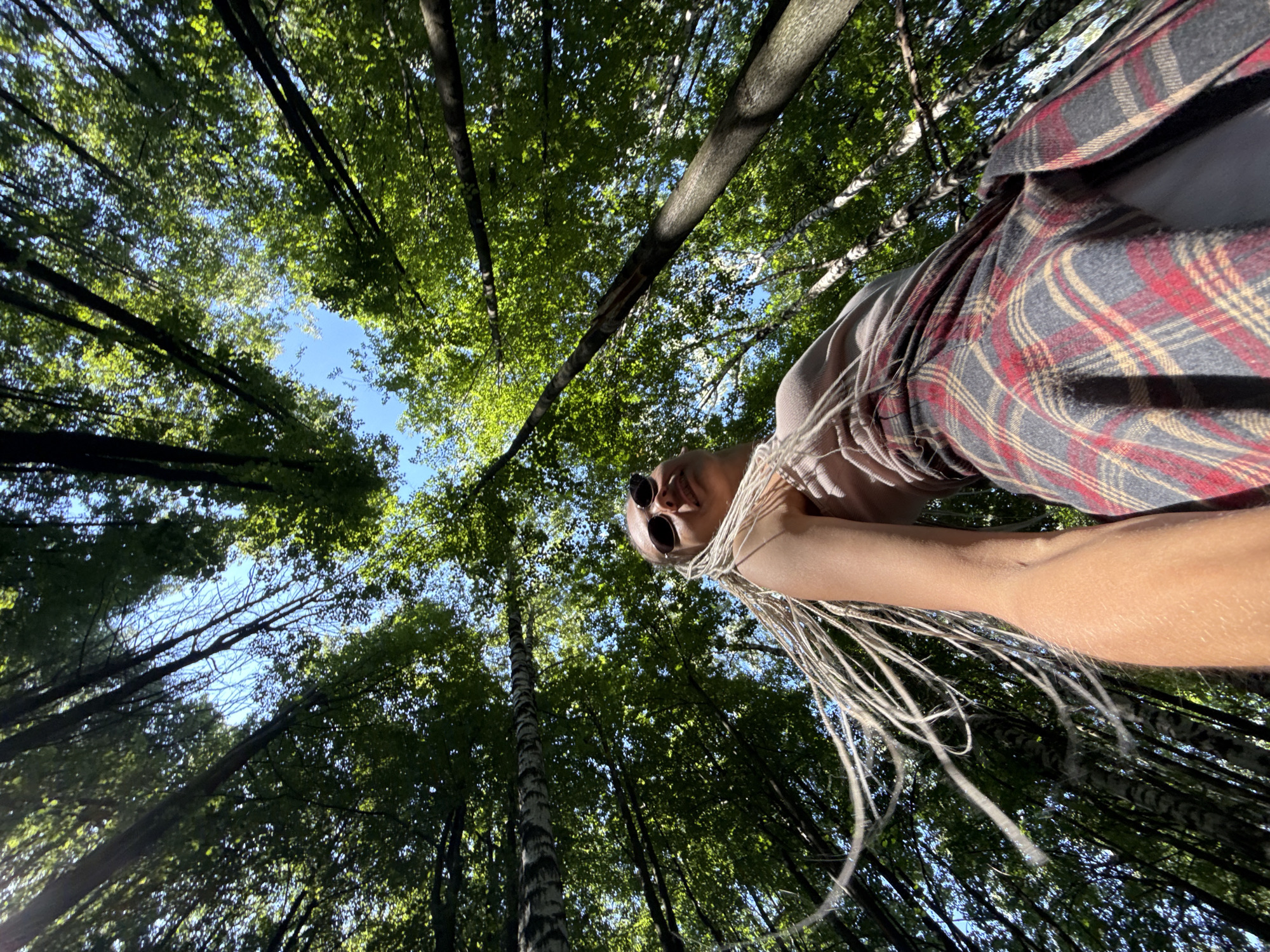 A low-angle shot captures a person with blonde dreadlocks wearing sunglasses and a plaid skirt in a forest. The person's head and upper torso are visible, with their hair flowing to the left of the frame. The background consists of a dense canopy of green trees and a clear blue sky, suggesting an outdoor setting during the daytime, possibly in Rusanovo, Russia. The person appears to be looking upwards, and the lighting indicates a sunny environment.