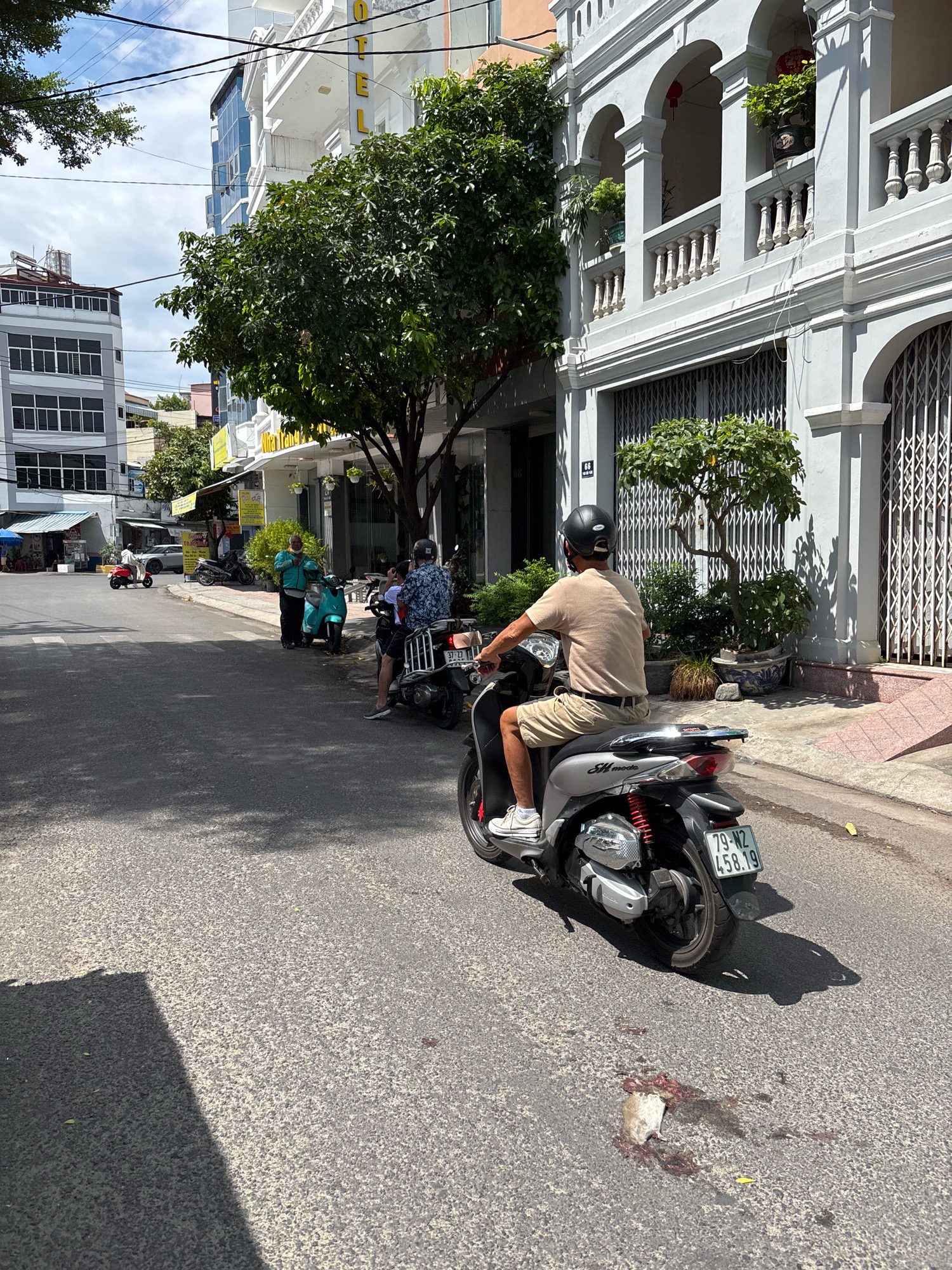 In Nha Trang, Vietnam, on a sunlit day, a man rides a silver scooter down a paved street. He wears a beige t-shirt, tan shorts, a black helmet, and white sneakers. The scooter has a license plate with the number 79-N2 458.19. In front of him, a dead rat lies on the asphalt, surrounded by blood. Buildings line the street, including a white building with arched balconies and a modern grey building. Several other parked scooters and a few people are visible in the background.