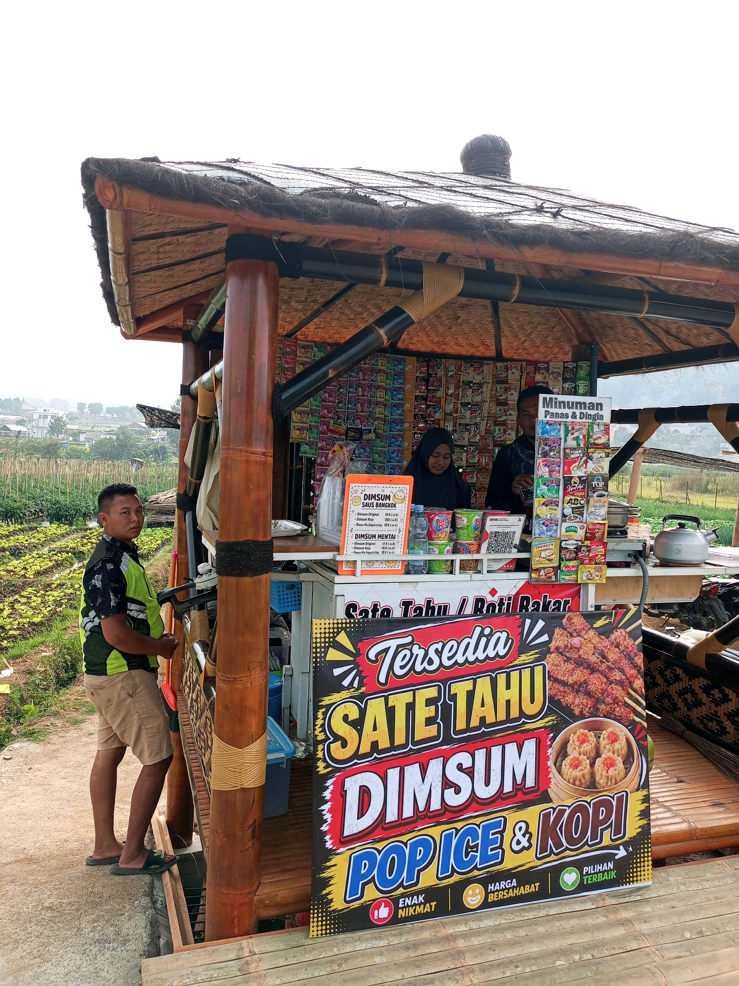 In Batu, Indonesia, a food stall offers satay, dim sum, pop ice, and coffee. A man stands to the left of the stall, wearing a lime green vest over a black and green patterned shirt and khaki shorts. Two vendors, a woman in a black hijab and a man, are visible inside the stall, attending to customers. The stall is constructed with bamboo and thatched roofing, creating a rustic ambiance. The background reveals lush green fields and distant buildings, suggesting a rural or semi-rural setting. A prominent sign advertises the available food and drinks.