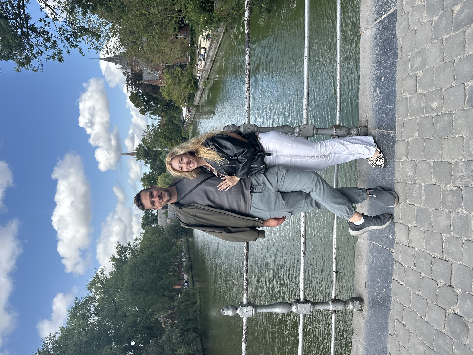 A medium shot shows a man and a woman standing on a stone bridge over a canal in Bruges, Belgium. The man is on the left, wearing a green jacket over a gray t-shirt and gray cargo pants. The woman is on the right, wearing a black leather jacket over a patterned top and white pants. They are standing close together, both smiling at the camera. The canal is wide and green, with trees and buildings lining its banks. In the background, a church spire is visible, along with more trees and a partly cloudy blue sky. The bridge has a metal railing with decorative posts. The pavement of the bridge is made of cobblestones.