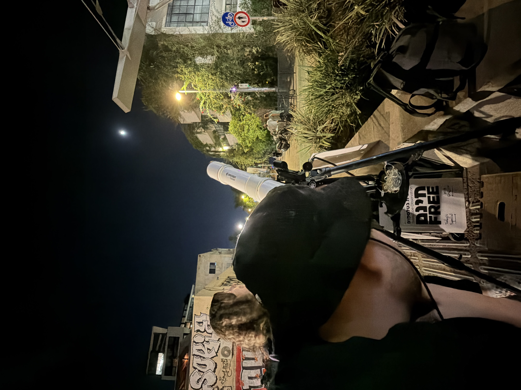 The media file shows a person wearing a black bucket hat, looking down at a telescope. The telescope is white and appears to be a refracting telescope, with the brand name "Bresser" visible. It is mounted on a tripod that is attached to a bicycle. The scene is set at night, with a bright moon visible in the dark sky. The background shows buildings with graffiti, trees, and streetlights, suggesting an urban environment. There are signs on the buildings and a traffic sign with a bicycle and pedestrian symbol. A backpack is also visible in the scene, placed on a ledge. The overall mood seems casual and perhaps exploratory, given the telescope. The visible text on the telescope reads "BRESSER". There is also text on a sign near the telescope, which includes "TODAY'S SPECIAL" and some Hebrew text. The location context indicates Tel-Aviv, Israel.