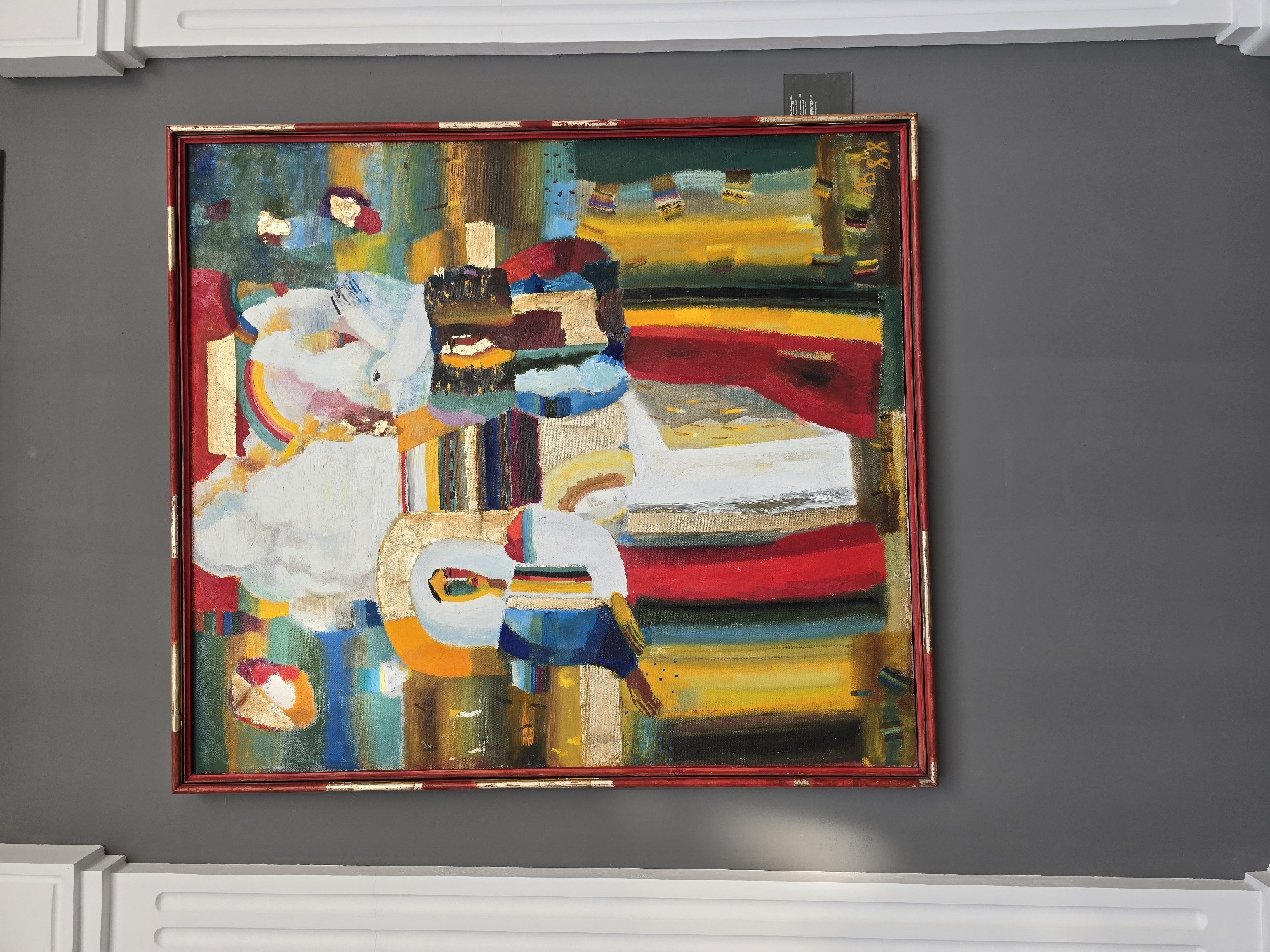 The image depicts a framed abstract painting with vibrant colors, hung on a dark gray wall between two white columns. The painting features two figures rendered in a stylized, geometric manner, with bold brushstrokes and a mix of bold colors like red, yellow, blue, and white. Gold leaf elements are incorporated into the artwork, adding a shimmering texture. A rainbow motif is subtly present in the upper portion of the painting. The bottom right corner of the painting shows a signature and the year "88". To the right of the painting, a small white plaque with black text is attached to the wall, likely providing information about the artwork. The lighting suggests an indoor exhibition space. The overall impression is one of a modern art display in a gallery or museum.