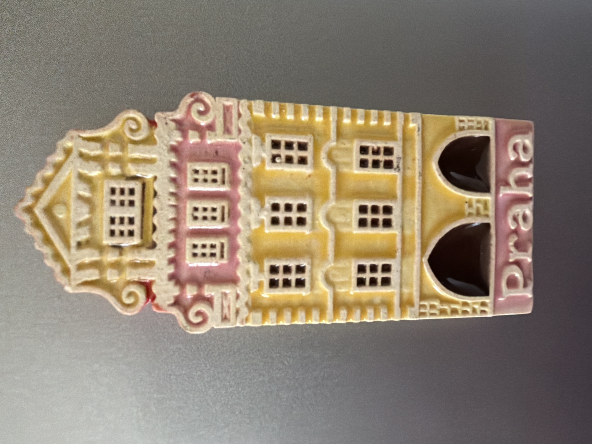 This is a close-up, eye-level, slightly angled, full-shot photograph of a decorative magnet shaped like a colorful, multi-story building. The magnet is pink, yellow, and cream-colored, with dark brown window details. The building has a stepped gable roof with scrolled decorative elements on the sides. There are at least three visible stories with windows, and arched openings at the bottom. The word "Praha" is embossed in cream letters on a pink base at the very bottom of the magnet. The magnet is attached to a metallic, grey surface, likely a refrigerator. The image does not depict the city of Pazardzhik, Bulgaria.