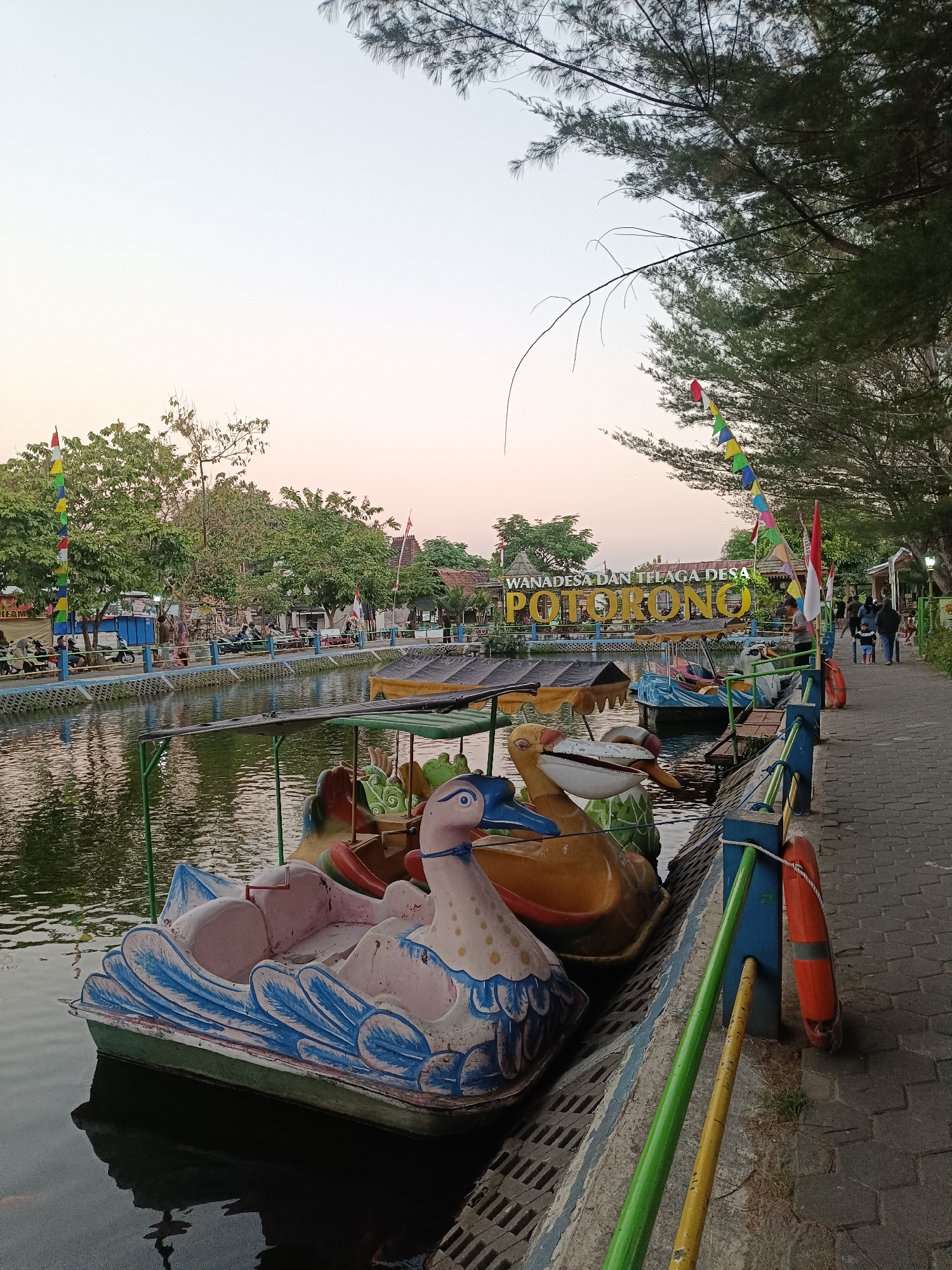 The image shows a canal-side walkway at Wana Desa dan Telaga Desa Potorono in Banguntapan, Indonesia. The walkway is paved with interlocking bricks and bordered by a blue railing with green and yellow poles. Several pedal boats shaped like animals, including a swan and ducks, are moored along the canal. In the background, trees line the canal, and a sign that reads "WANA DESA DAN TELAGA DESA POTORONO" is visible. A few people are walking along the path. The sky is a light, hazy blue, suggesting it is late afternoon or early evening. The water in the canal is murky and reflects the surrounding environment.