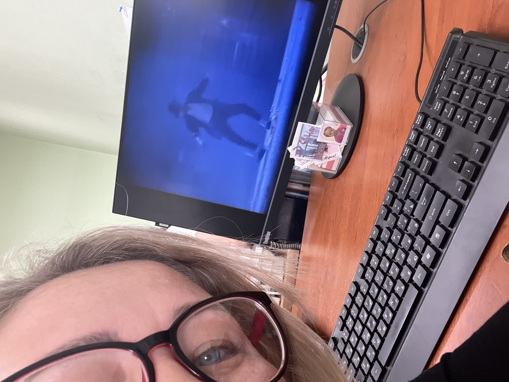 The image shows a close-up selfie of a person wearing glasses, with a computer monitor and keyboard in the background. The monitor displays a blue-toned scene with a figure silhouetted against it. To the right of the monitor is a small, clear plastic container holding cards or photographs, one of which has text in Russian: "8 Mapmal," referring to "8 March." The black keyboard is a standard size and has Russian letters. The desk surface is a dark reddish-brown. The person's eye is blue. There are no indications of the location of this image, therefore it is impossible to place it in the city of Chaikovsky.
