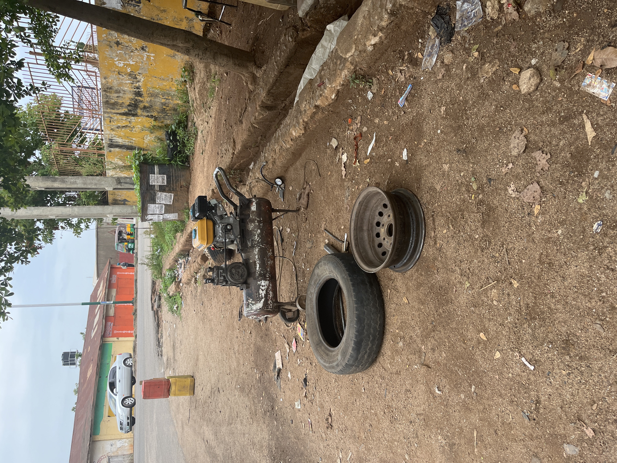 The image depicts an outdoor tire repair shop located in Jos, Nigeria. A tire and a wheel rim are on the ground, alongside an air compressor. The ground is dirt and littered with debris. In the background, there is a street with a car, a building with red and green accents, and a tricycle taxi. A tree and weathered buildings are also visible.