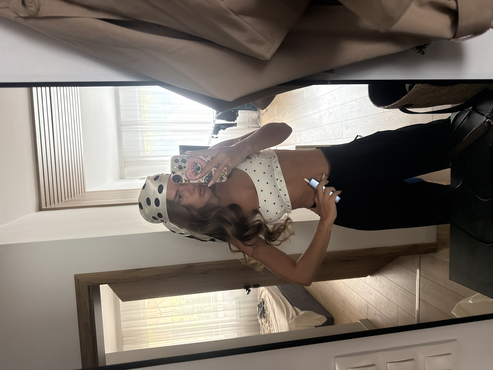 The image shows a woman in a white polka dot bikini top and black pants, taking a mirror selfie. She is wearing a polka dot headscarf. She is holding a phone in her right hand and a small white object in her left hand. The background shows a bedroom with a bed and a closet, and a doorway leading to another room. The scene appears to be indoors, possibly in an apartment or hotel room. There is no information to suggest this image is located in Plovdiv, Bulgaria.
