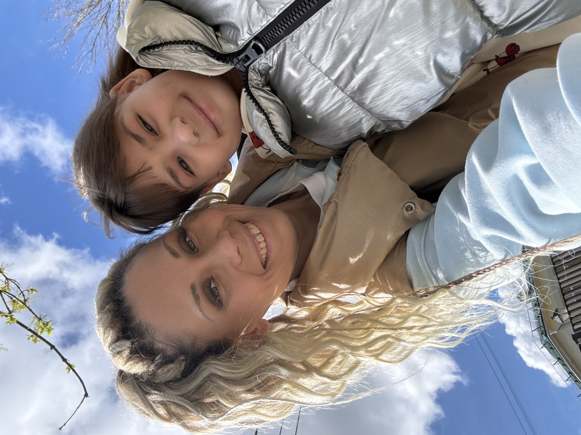 A woman and child are in a close embrace, likely a selfie taken from a low angle. The woman has blonde hair, styled back and braided. She is smiling broadly. The child, with brown hair, also smiles. Both appear to be wearing jackets. The background shows a bright blue sky with puffy white clouds. A tree branch is visible on the left side and a building structure on the right. Given the location information provided, this photo was likely taken in or near Rusanovo, Russia.