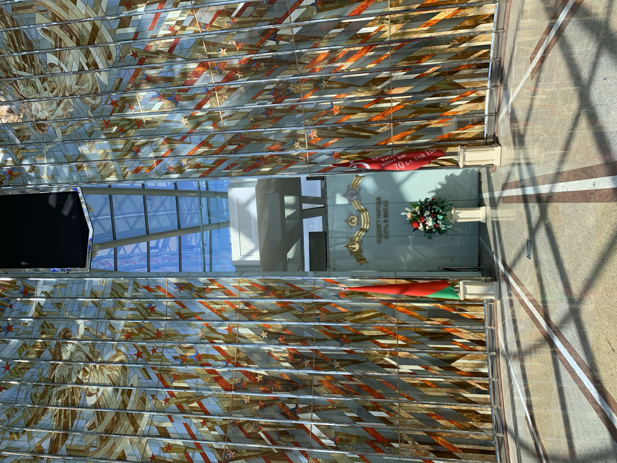 This indoor shot features a detailed mosaic artwork and a memorial display in what is likely a public space. The mosaic spans a wall and possibly a ceiling, showing figures and geometric designs in shades of blue, orange, brown, and beige. In front of the mosaic, there's a glass display case with military-related memorabilia including a text inscription: "ПОДВИГУ НАРОДА ЖИТЬ В ВЕКАХ," which translates to "To the people's feat to live for centuries" and the words "KUTUSU ИДРИЦК. ДИВ. 70 с.к. 34.д. 15.9.", likely referring to a military unit. Red and green ribbons or flags are present. A vase of flowers sits below the display. The floor is marble with geometric patterns. The location is strongly indicated to be in Minsk, Belarus, given the language and symbolism within the display.