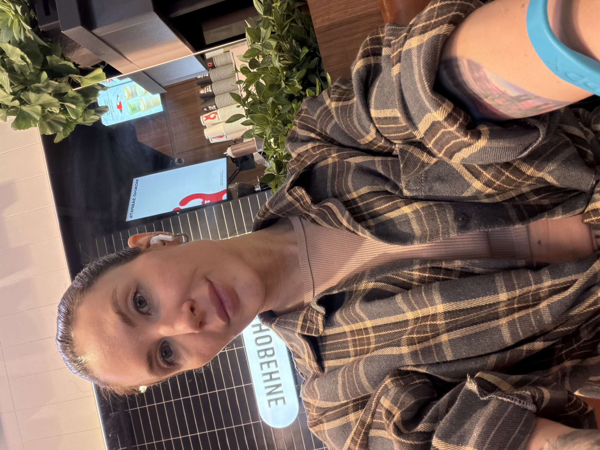 A woman with gray eyes and dark hair pulled back is taking a selfie. She wears a plaid shirt over a light pink top. A blue fitness tracker is on her arm. There is a tattoo on her arm. She is seated in what appears to be a cafe or restaurant. Behind her, there is a sign in Russian, "HOBEHHE," indicating the location is likely in Russia. In the background, there is a display with food and drink items, and plants. The location is the city of Perm, Russia.