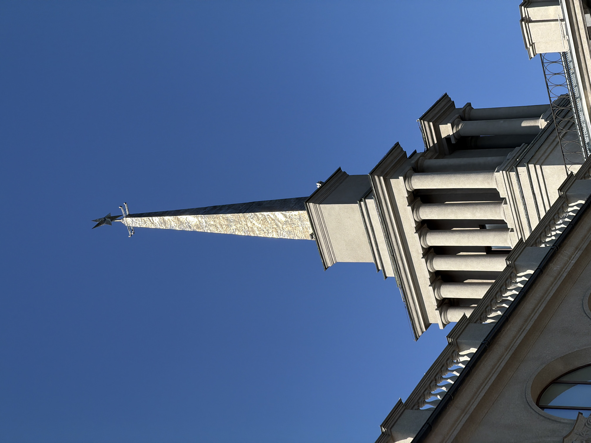 The image shows a low-angle view of a building against a clear blue sky. The architectural features include a spire with a star at the top, classical columns, and decorative railings. The building is the Krasnoyarsk Regional Local Lore Museum, located in Krasnoyarsk, Russia.