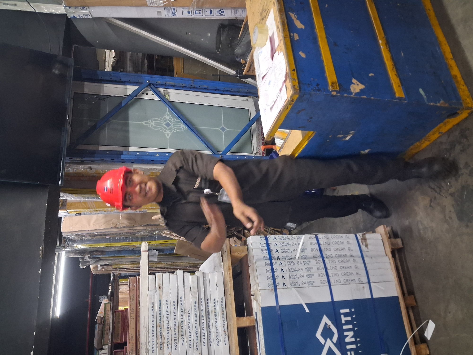A man wearing a red hard hat and a dark uniform stands in a warehouse in Depok, Indonesia. He is facing the camera and smiling, with his hands raised as if gesturing or explaining something. In the background, stacks of tiles and building materials are visible, along with shelving units and a blue counter with papers and tape on it. The lighting suggests an indoor industrial setting.