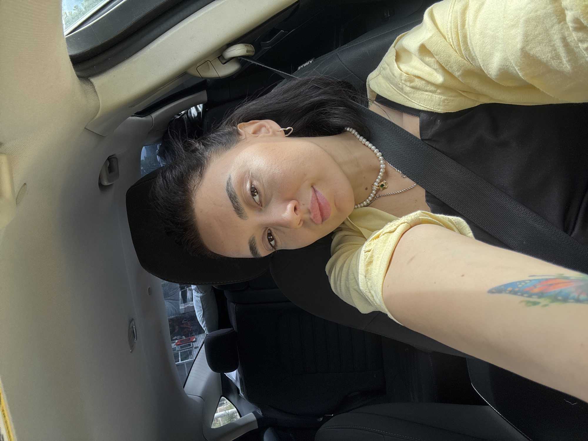 A young woman with dark hair is sitting in the driver's seat of a car. She is wearing a yellow shirt over a black top and a pearl necklace with a pendant. She has a tattoo on her forearm. The car's interior is black. The background visible through the car window suggests an urban environment. The exact location in Tbilisi, Georgia, cannot be determined from the image.