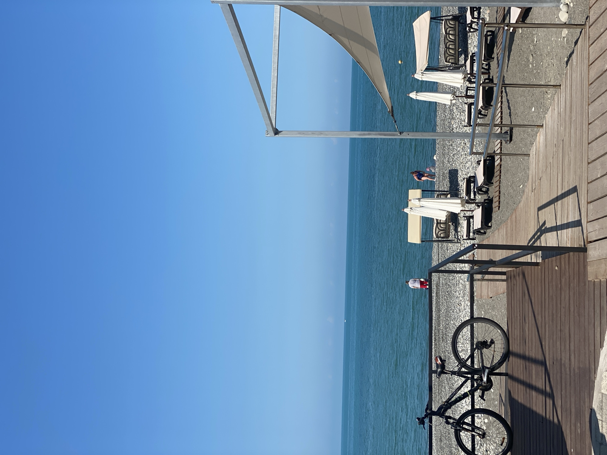 A daytime, medium shot captures a scene on a pebble beach in Sochi, Russia. A black bicycle is parked on a wooden boardwalk that leads down to the beach. The boardwalk is bordered by a metal railing. Beyond the railing, the pebble beach is dotted with lounge chairs and closed white umbrellas, some of which are arranged around a small gazebo-like structure. The turquoise sea stretches to the horizon under a clear blue sky. Two people are visible near the water's edge. One person, wearing a striped shirt and red shorts, stands facing away from the camera. Another person in a blue swimsuit is partially visible with a small child. The overall atmosphere is one of a quiet, sunny day at the seaside.